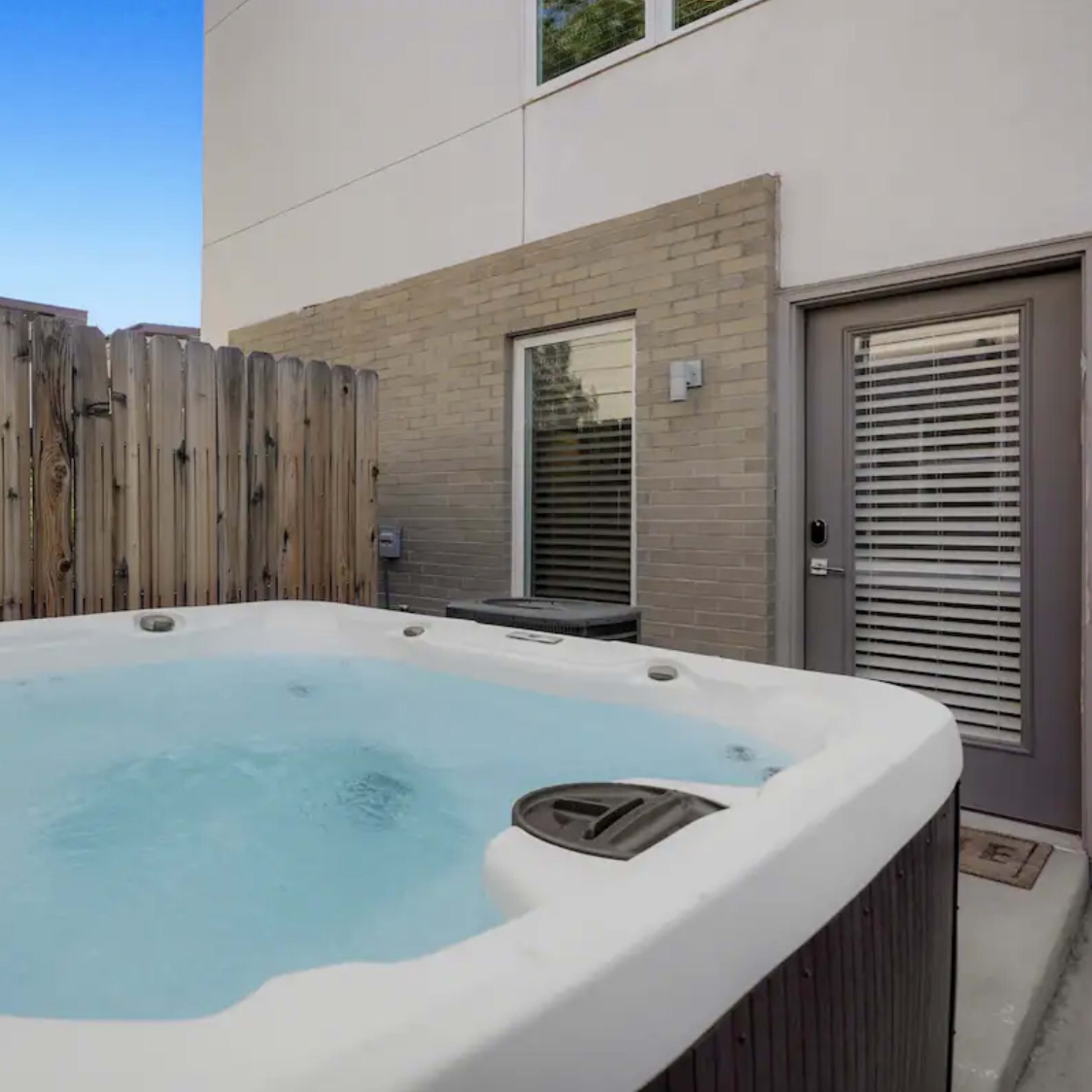 A hot tub filled with water is located in a small outdoor area surrounded by a wooden fence and adjacent to a modern building.