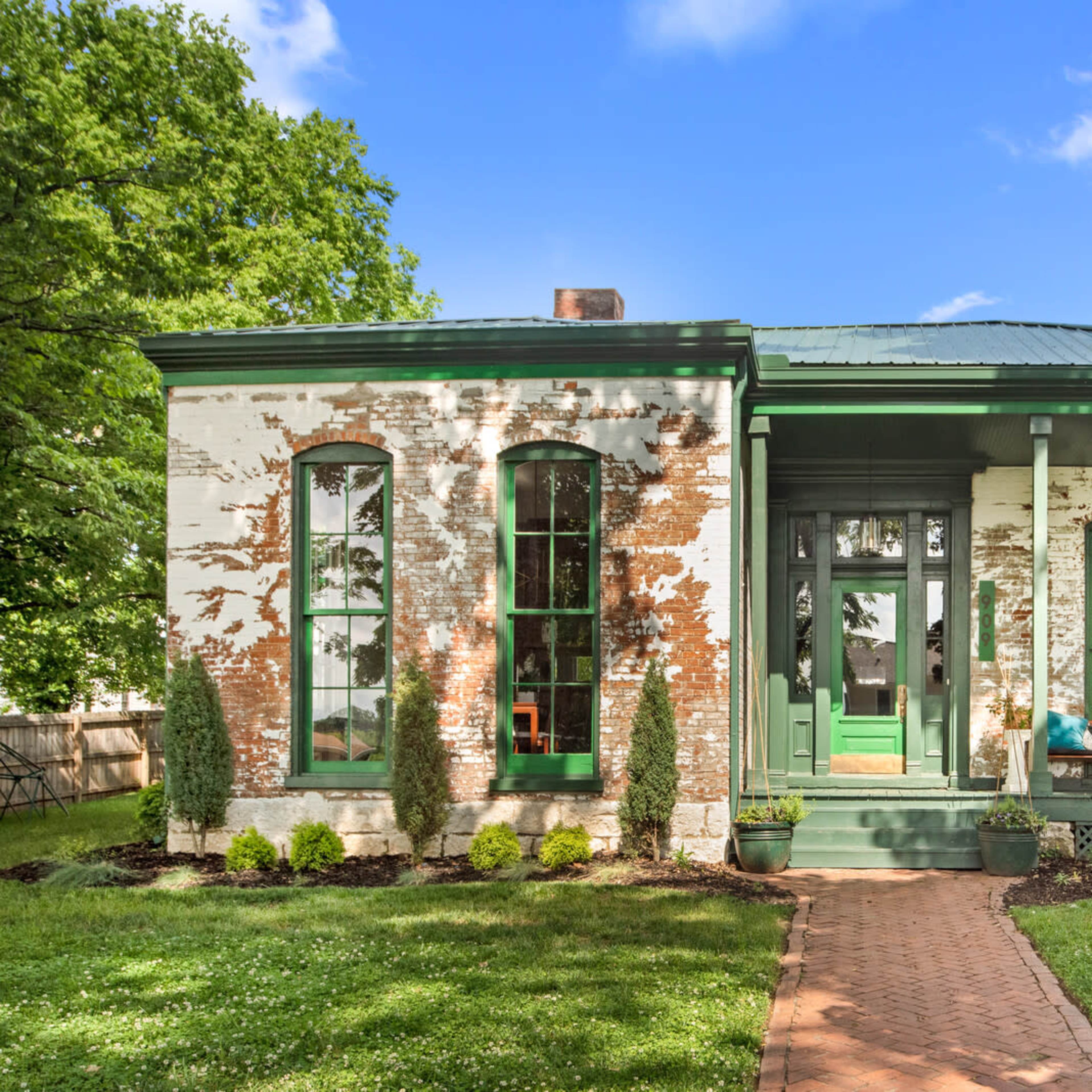 A historic house with a green trim, featuring exposed brick walls and a front porch, surrounded by manicured grass and shrubs.