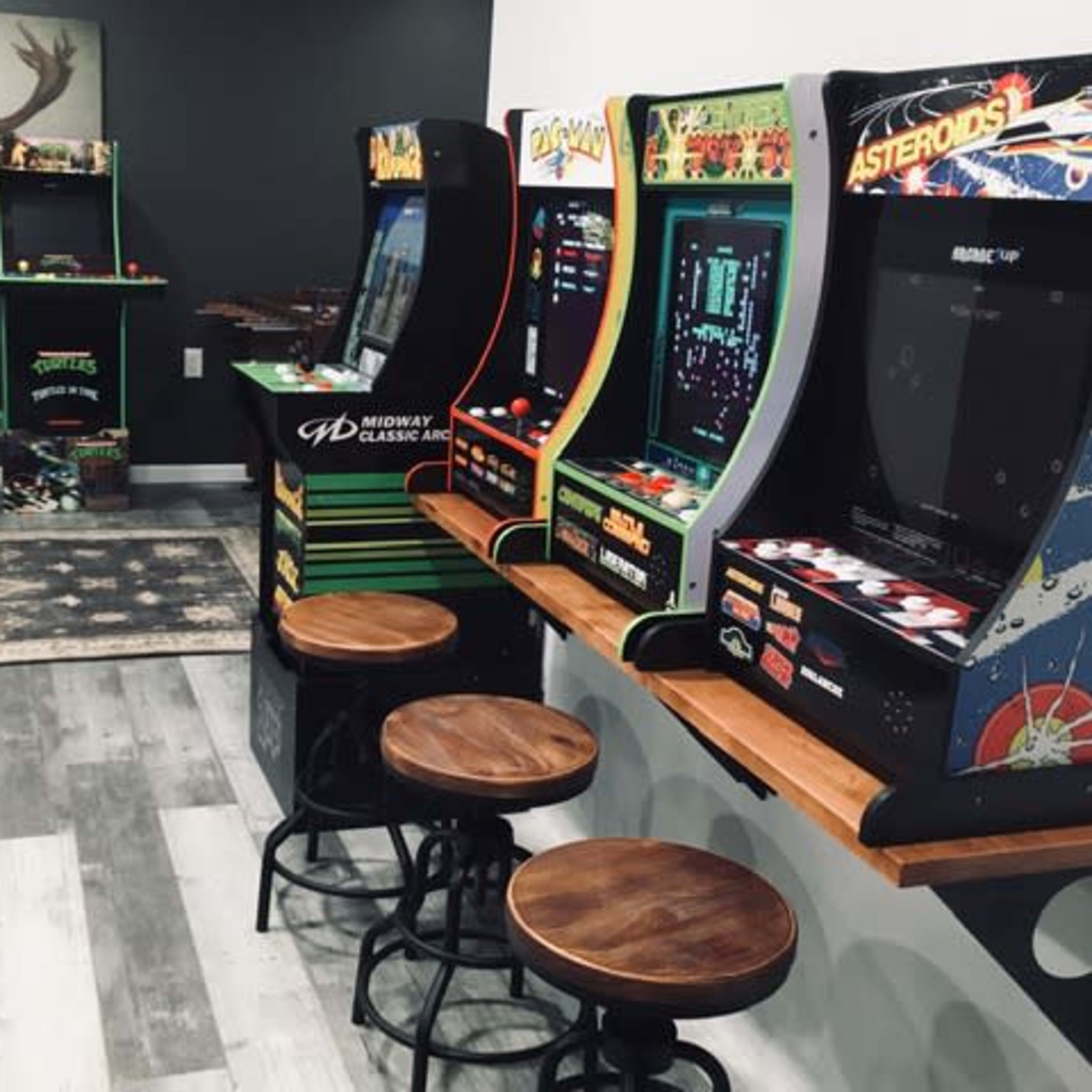 A game room featuring multiple retro arcade machines lined up along one wall, accompanied by wooden stools for seating.