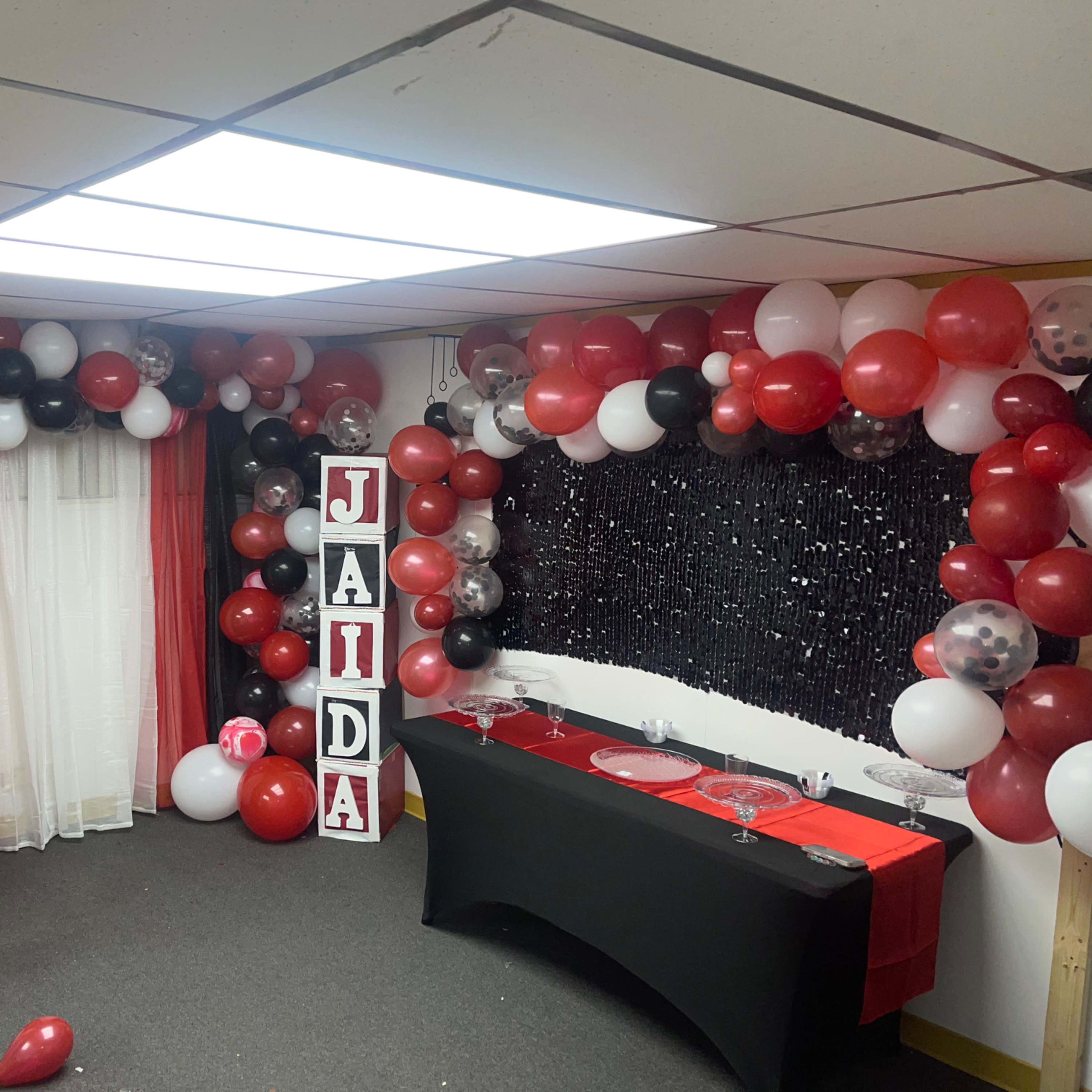 A decorated party space with red, black, and white balloons, large letters spelling "JALEYAH" and "JAIDA," and a table set up with a black tablecloth in front of a glittery backdrop.