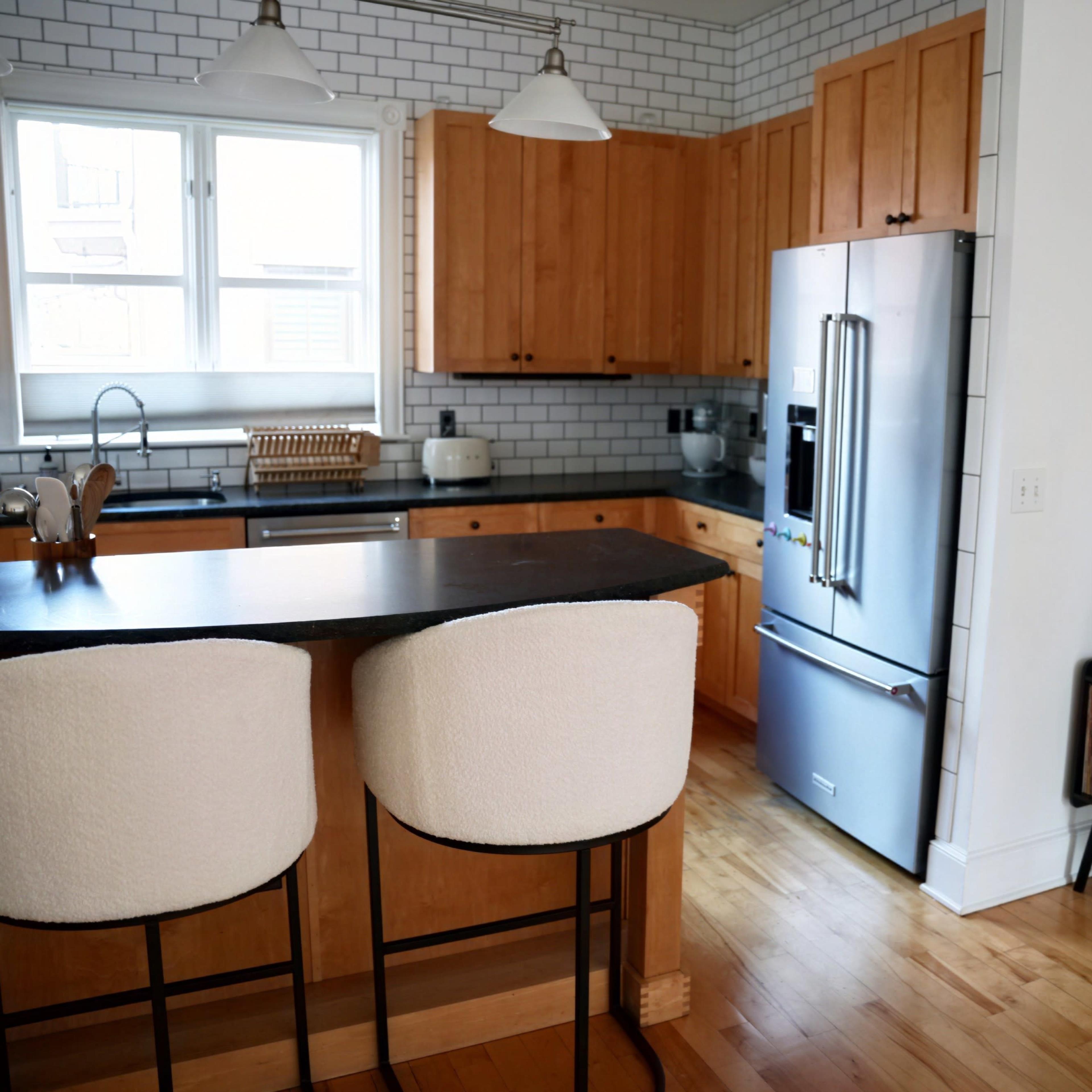 A modern kitchen with wooden cabinets, a black countertop, and a refrigerator, alongside a seating area with two bar stools and a vintage record player in the corner.