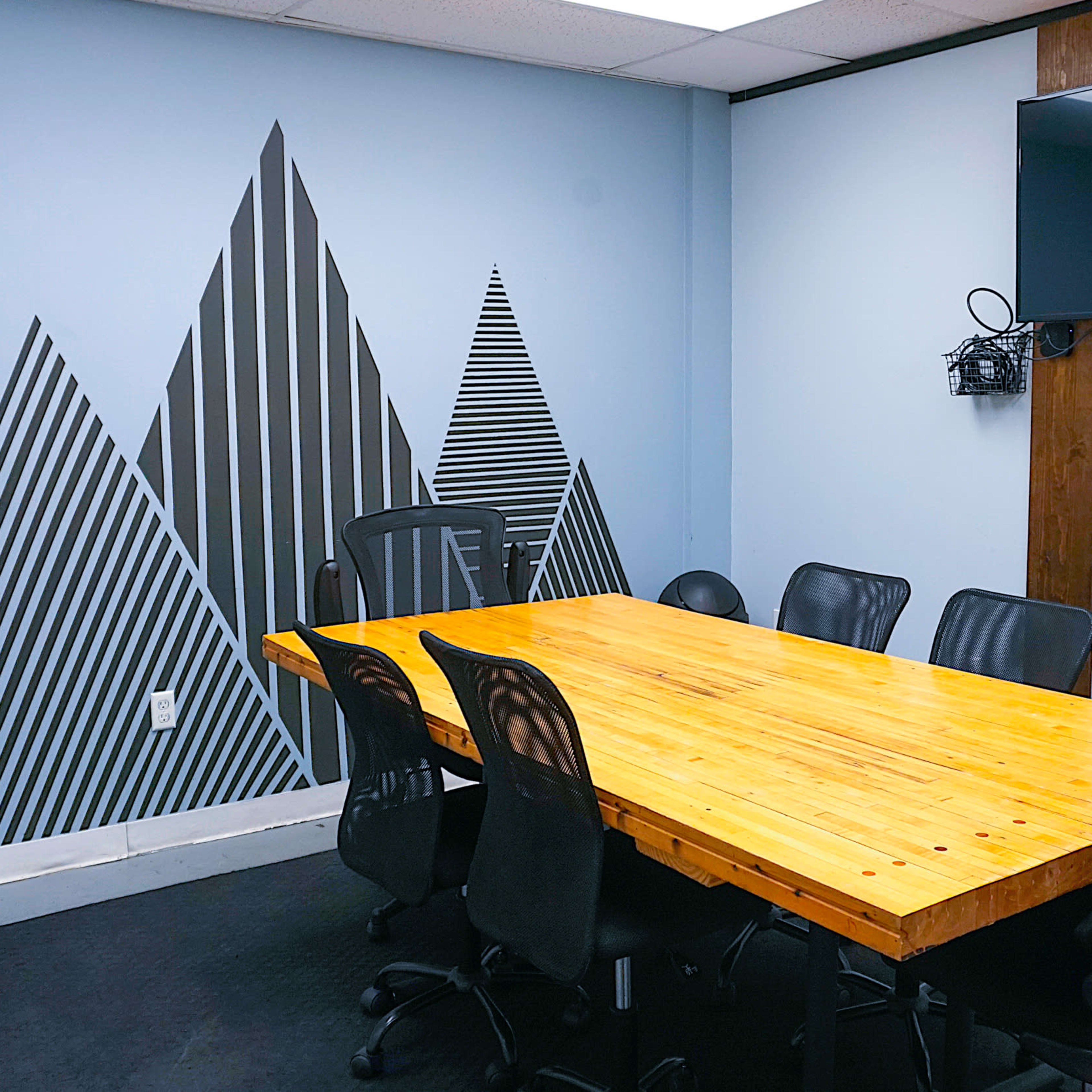 A conference room with a large wooden table surrounded by black chairs, featuring a wall decoration of striped mountains and a mounted television.