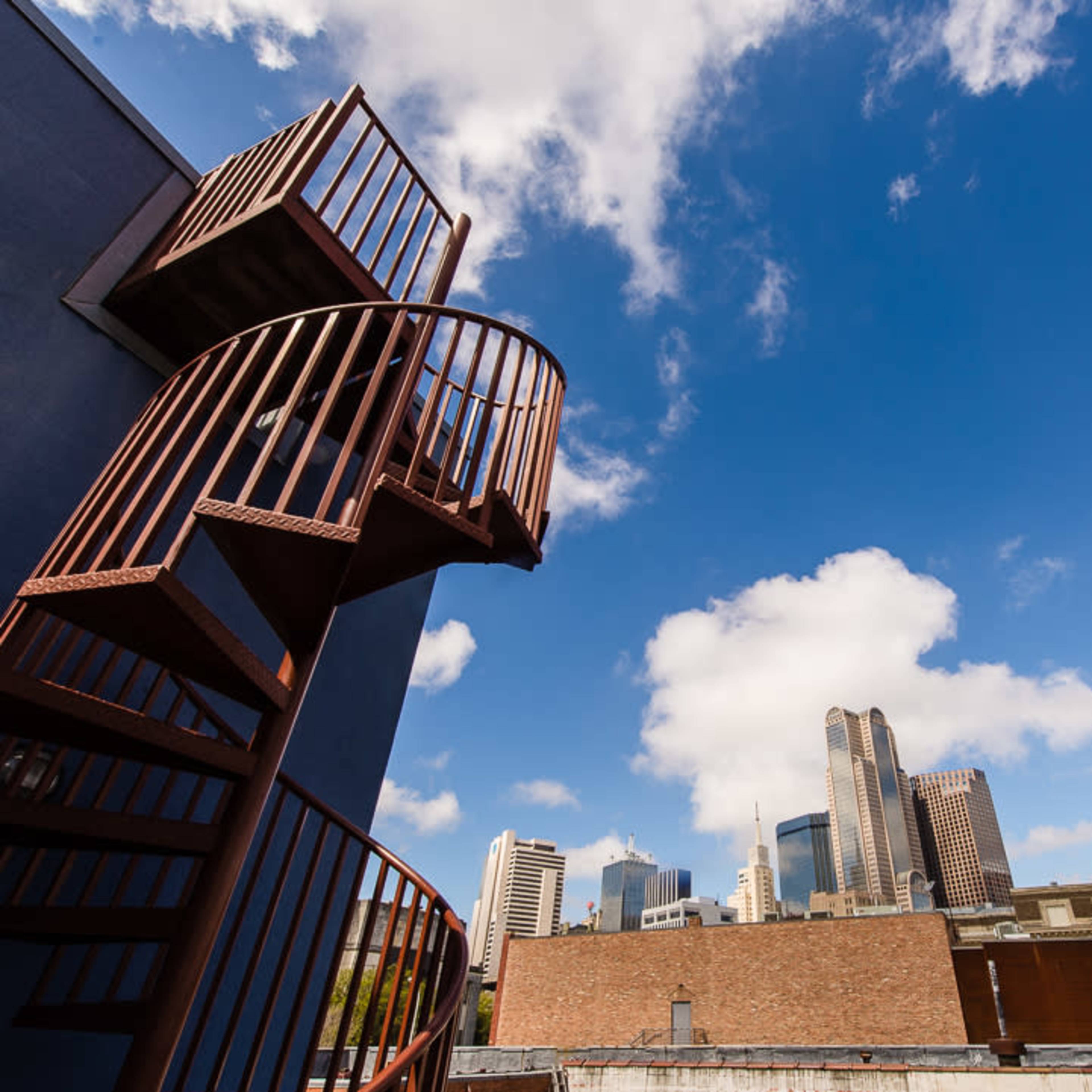 A spiral staircase ascends from a rooftop, leading to a view of a city skyline under a partly cloudy sky.