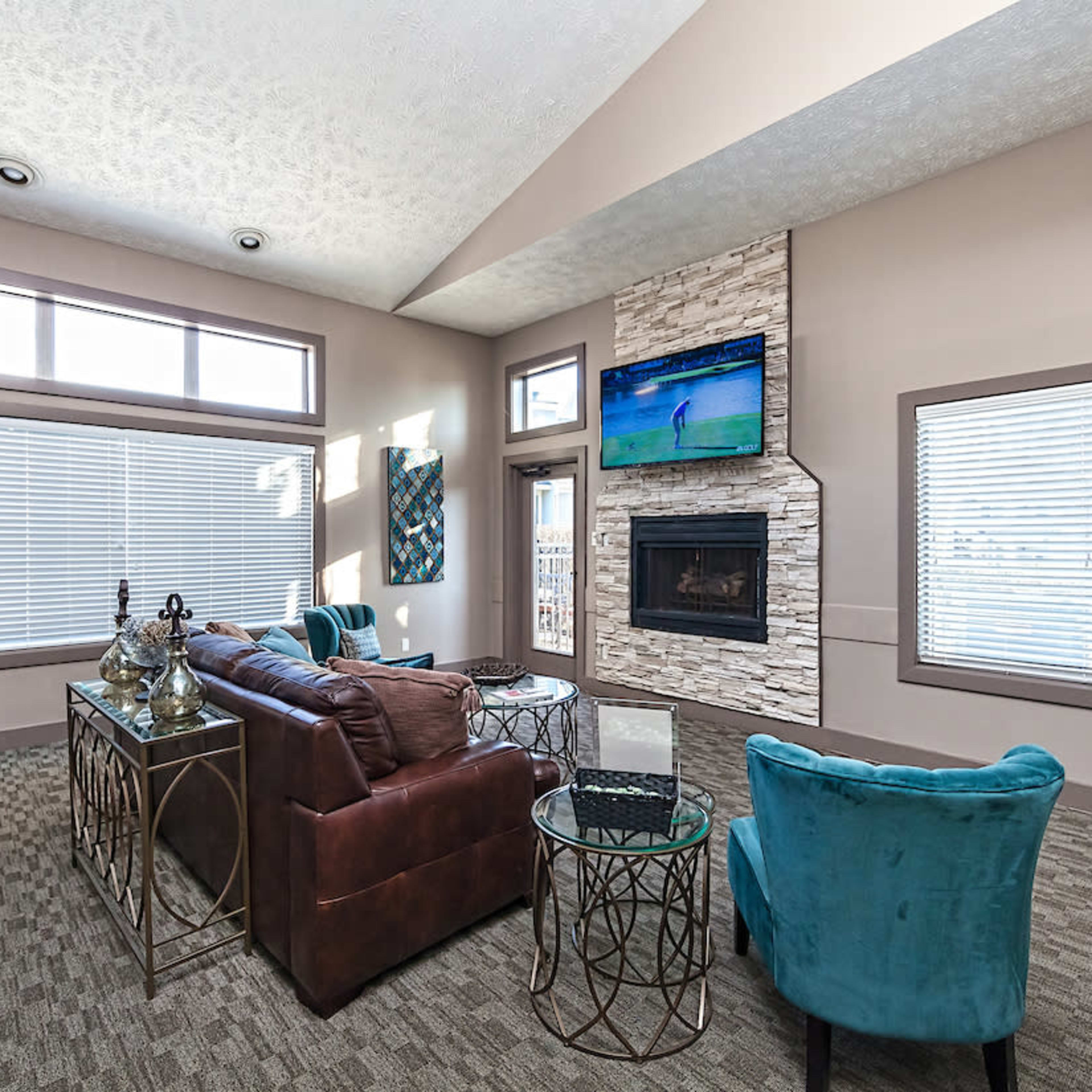 A modern living room with a brown leather sofa, teal accent chairs, a stone fireplace, and large windows allowing natural light to enter.