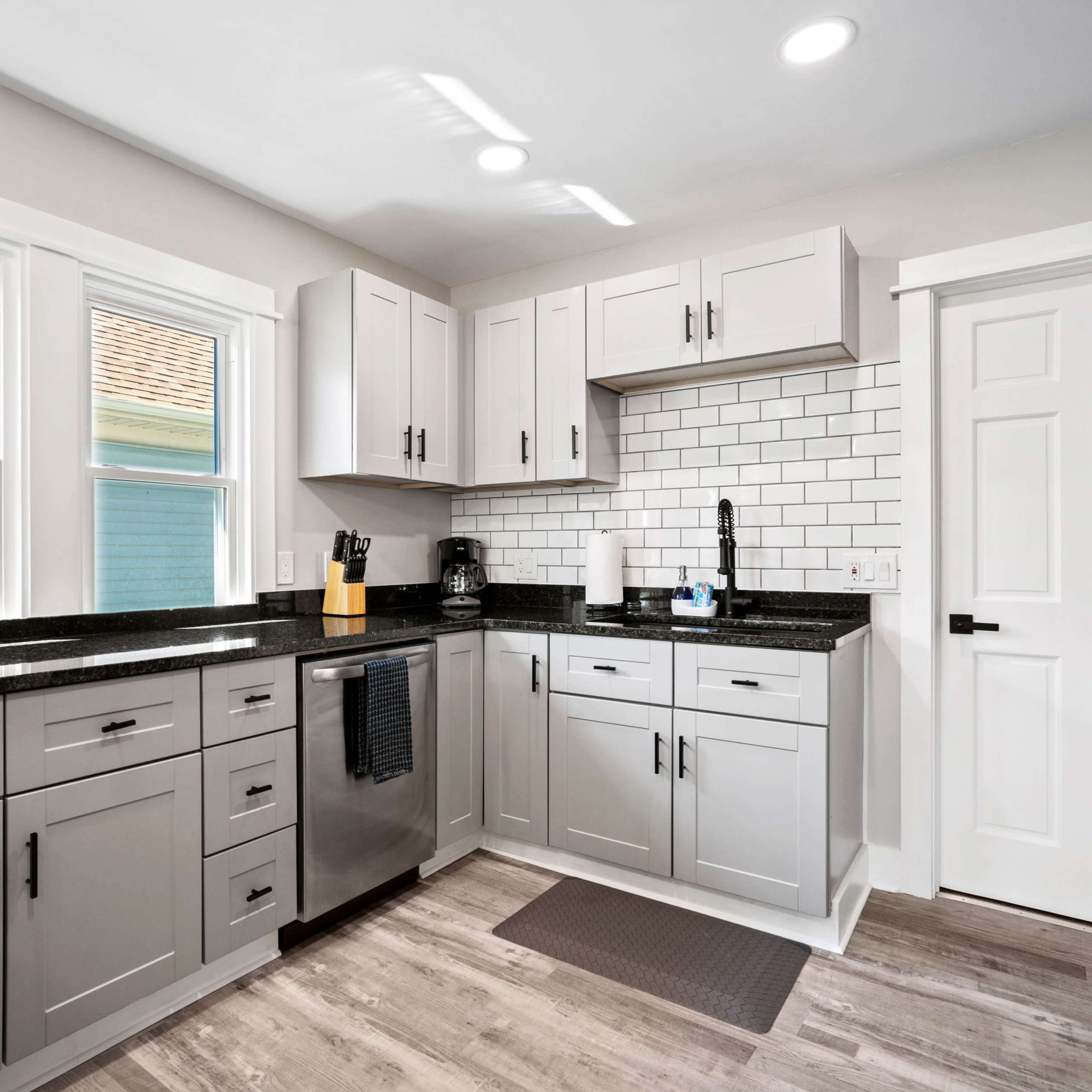 A modern kitchen features gray cabinets, black countertops, and white tile backsplash with a window and door for natural light.