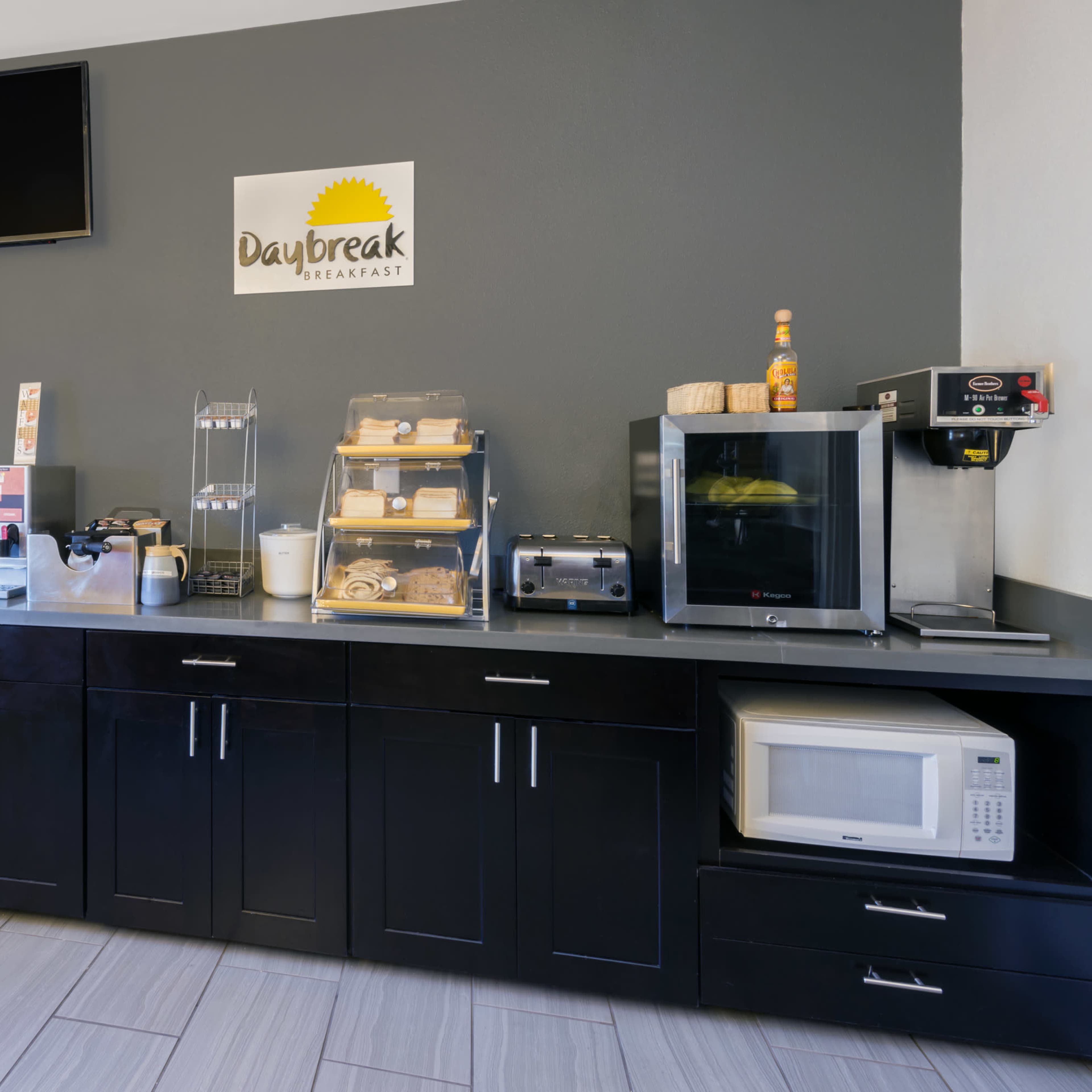 A breakfast area features a countertop with various appliances, including a coffee machine, toaster, microwave, and food display cases, against a gray wall.