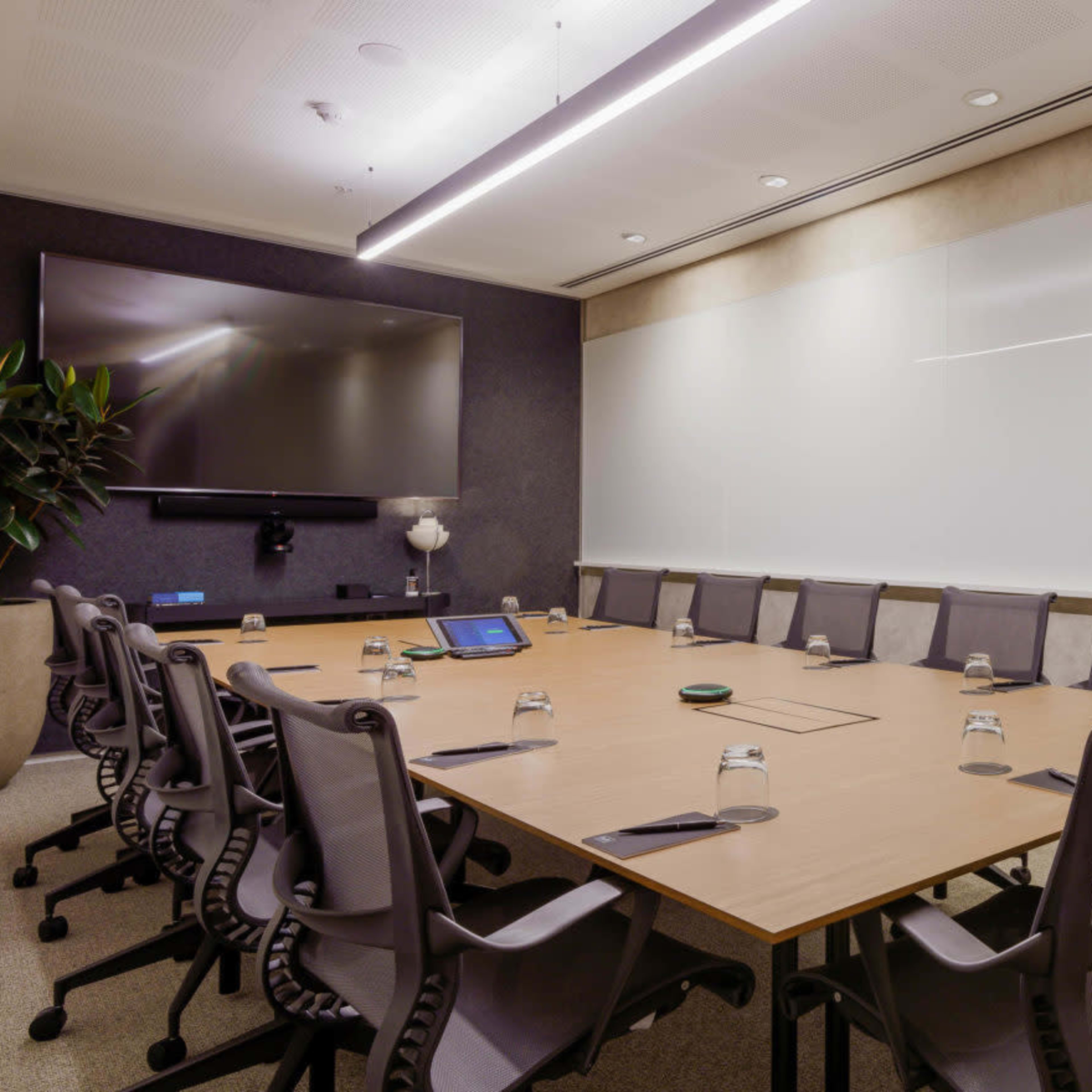 A modern conference room features a large table surrounded by ergonomic chairs, a wall-mounted television, and a whiteboard.
