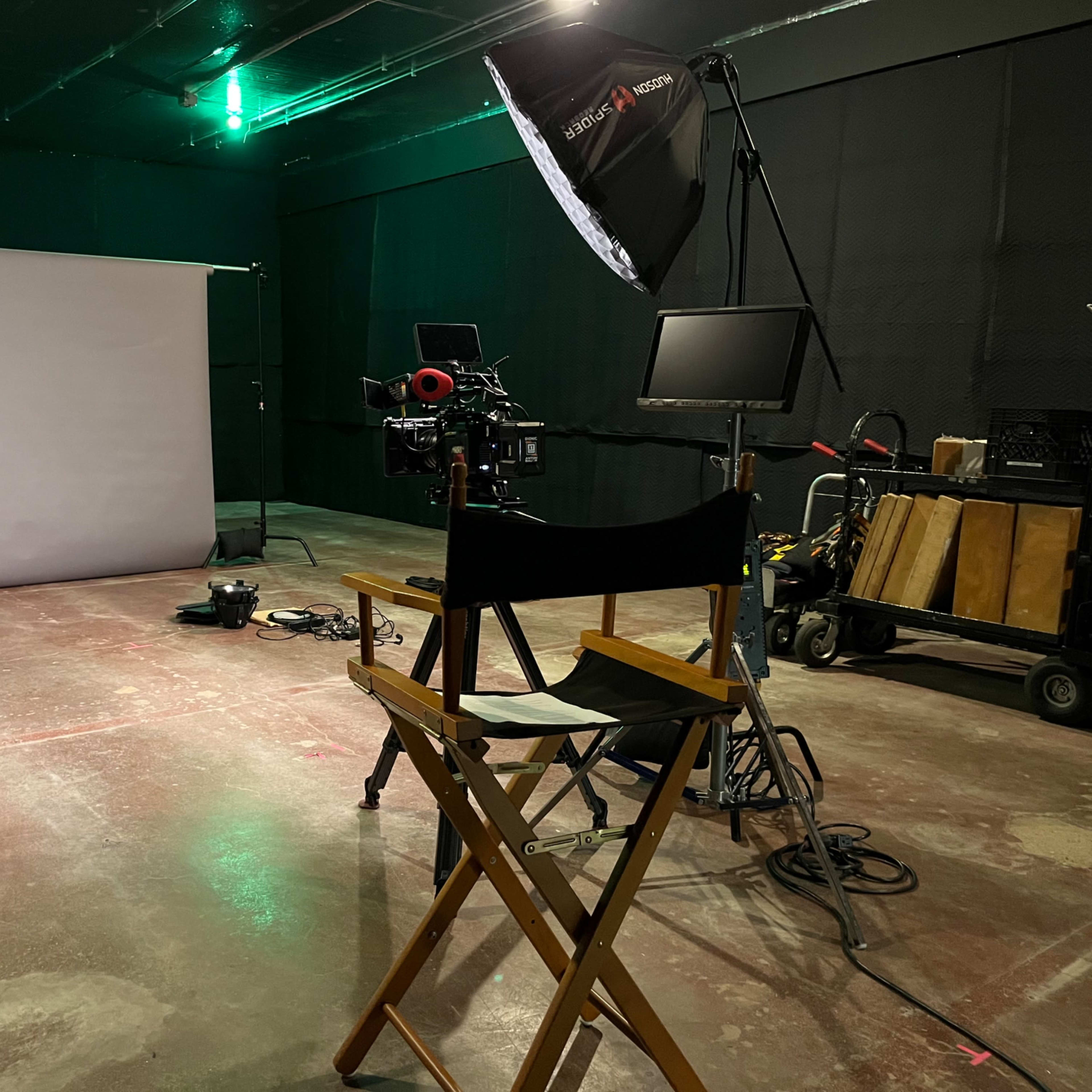 A film set featuring a camera on a tripod, a light umbrella, a monitor, and a director's chair in front of a gray backdrop.