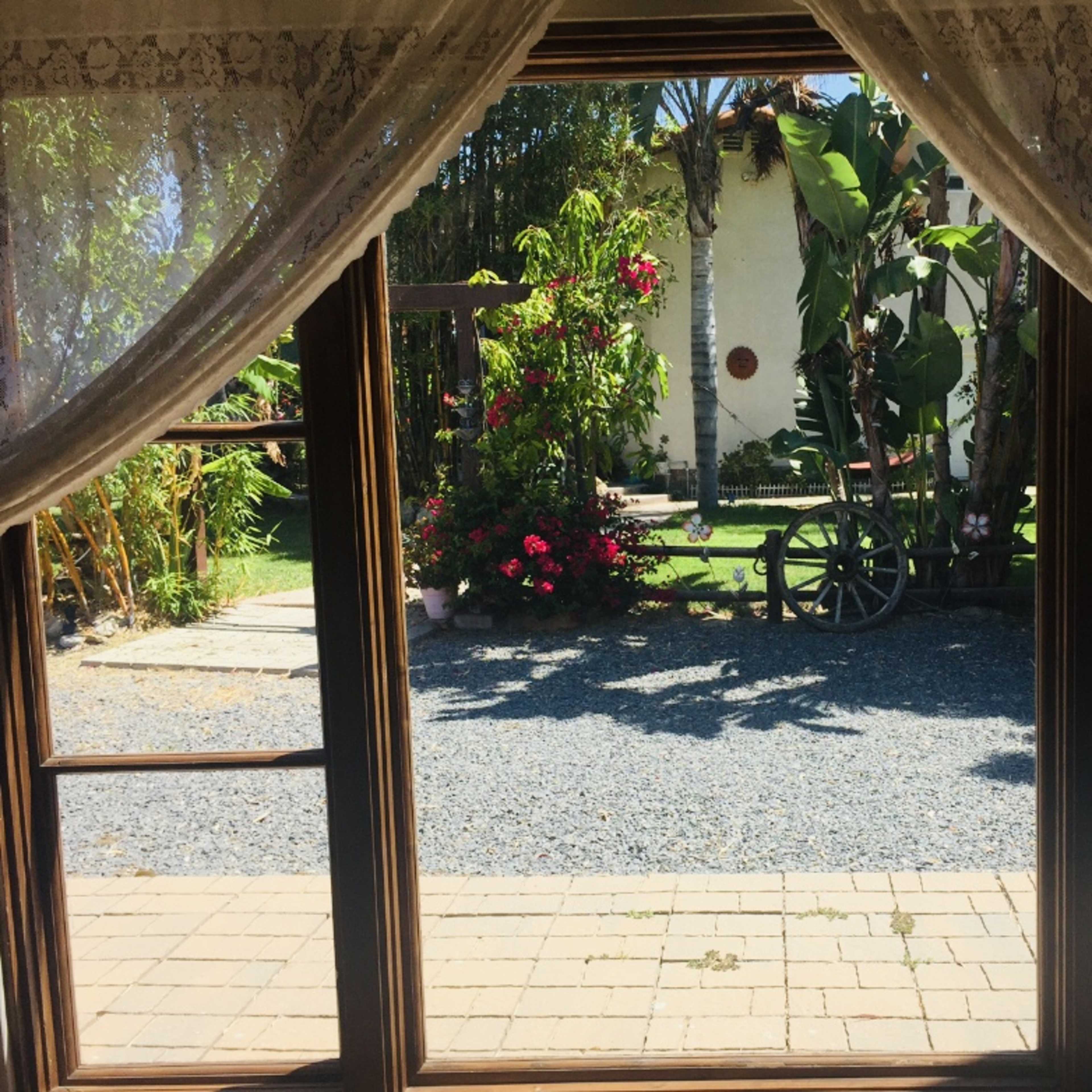 The view through the lace curtains shows a garden with a stone path, vibrant flowers, and a wooden wagon wheel.