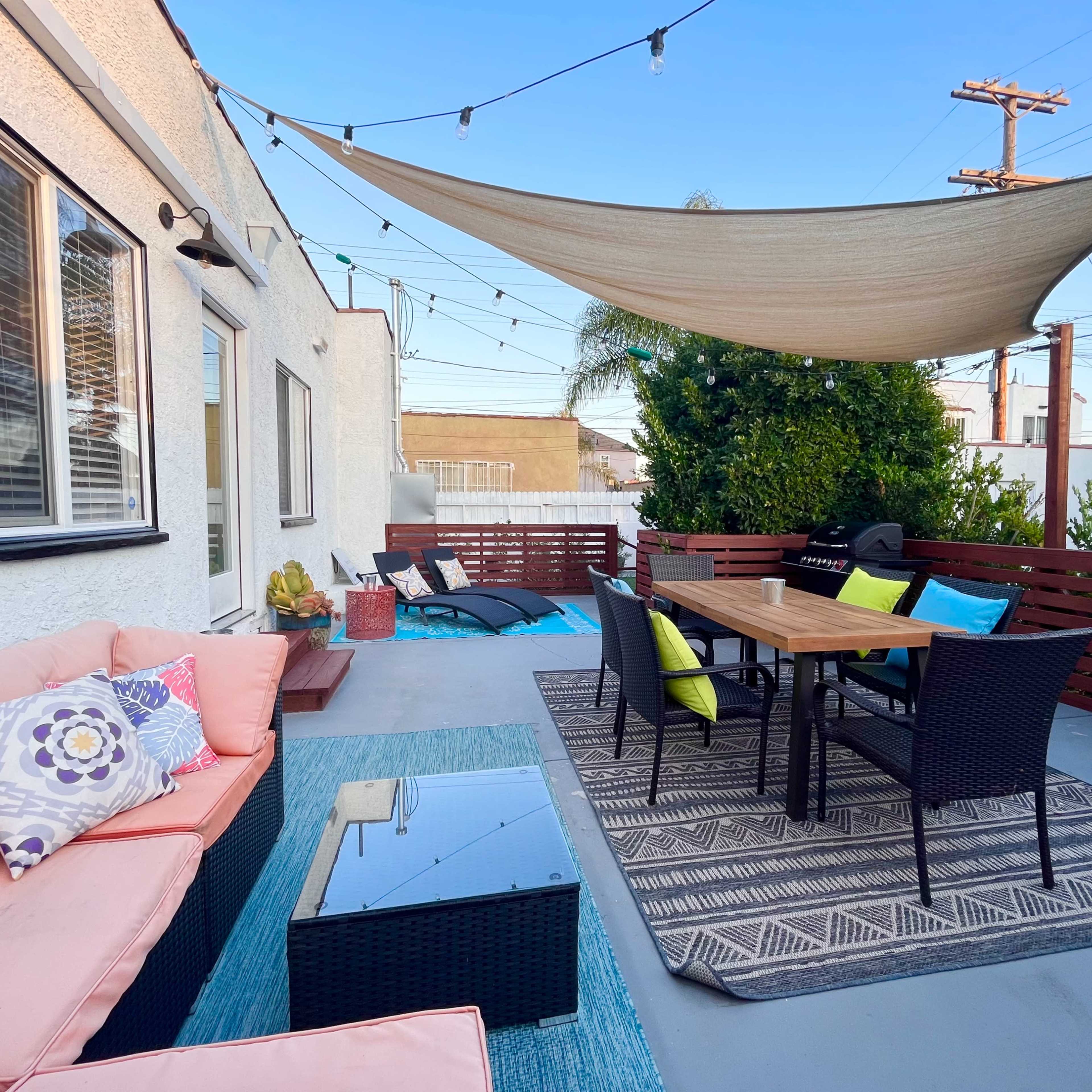 A patio with a dining table, black wicker chairs, a couch with colorful cushions, and a tarp overhead, surrounded by greenery and string lights.