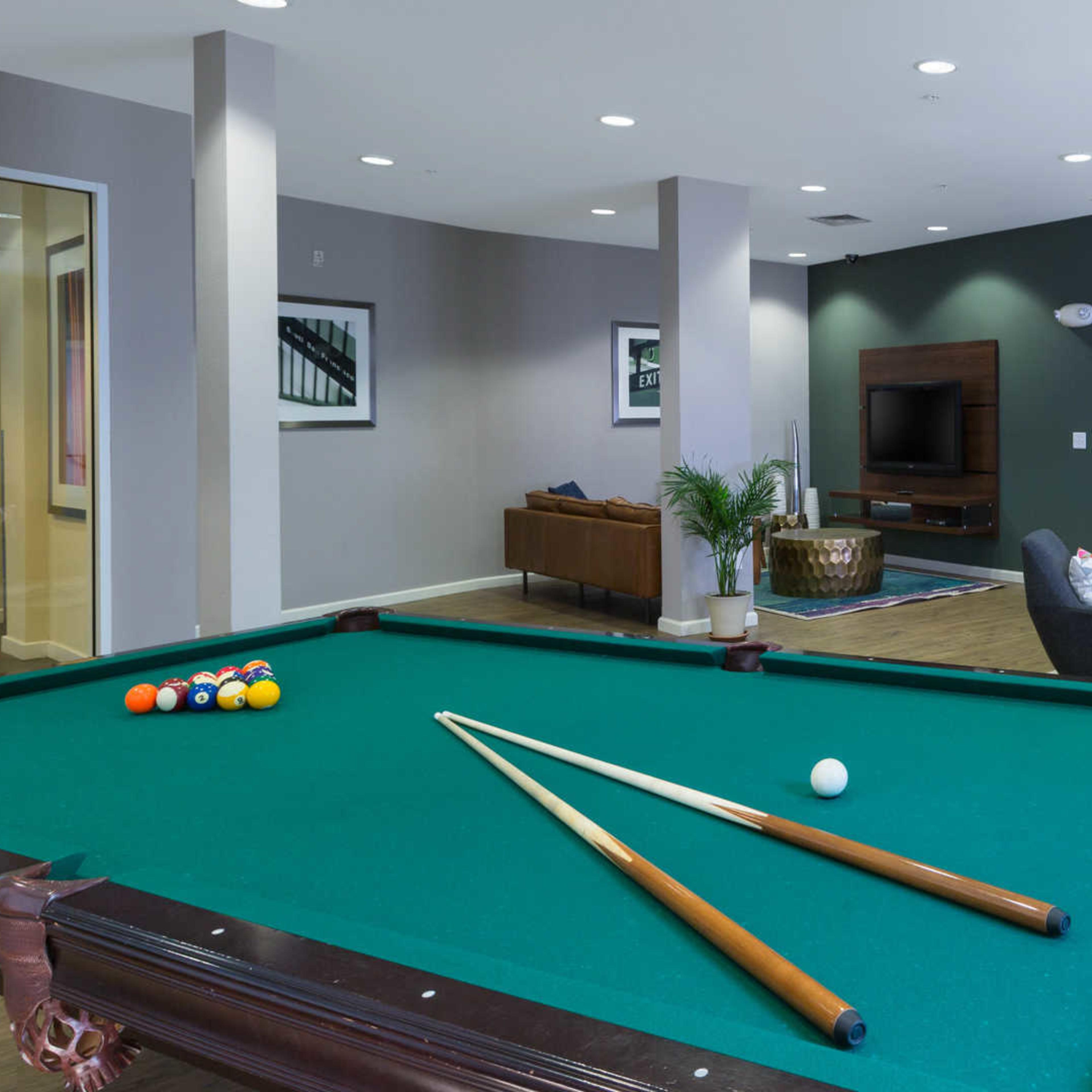 A green pool table with two cue sticks is prominently featured in a modern lounge area that includes seating and a television.