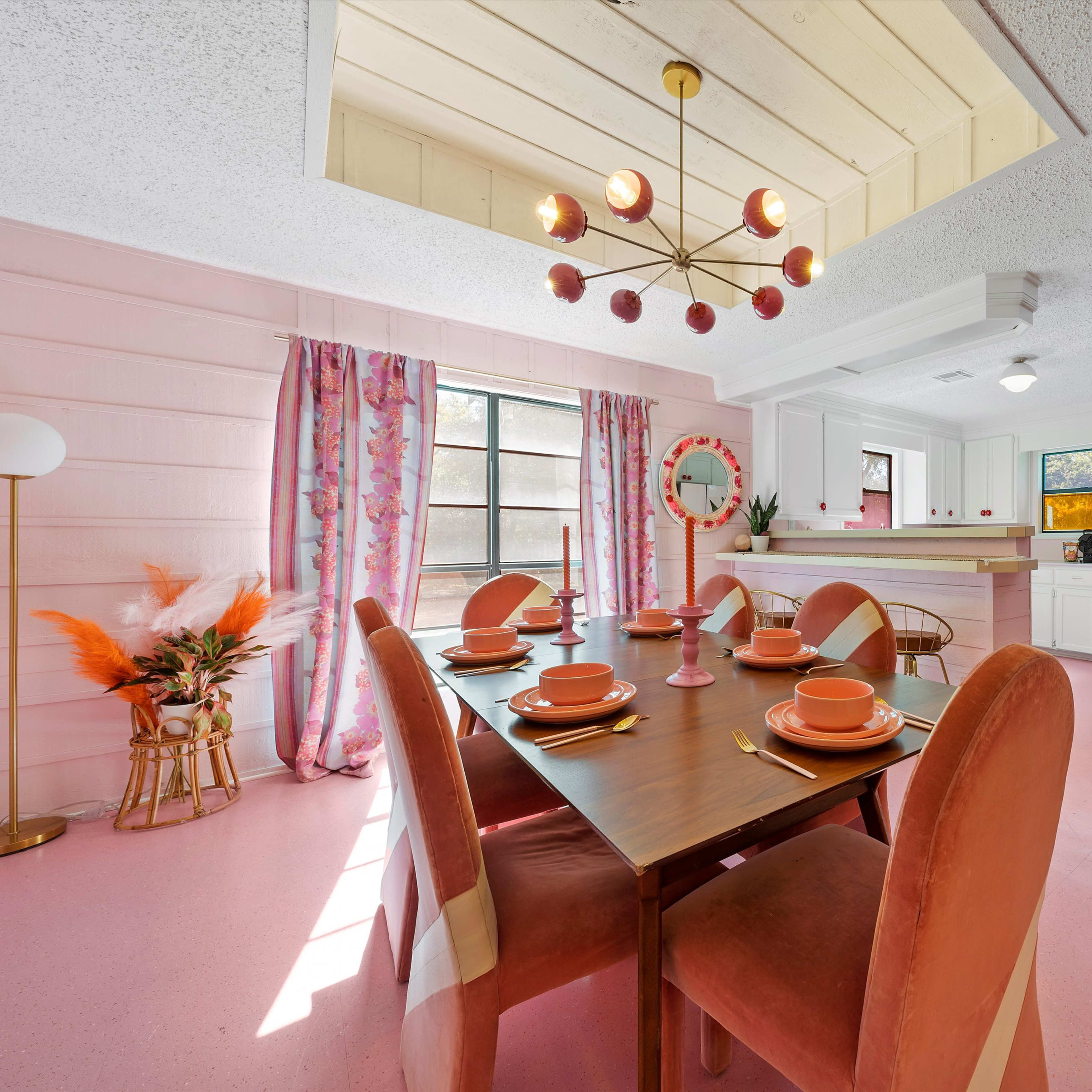 A brightly colored kitchen and dining area with a pink floor, a table set for dinner, and decorative elements like curtains and a chandelier.