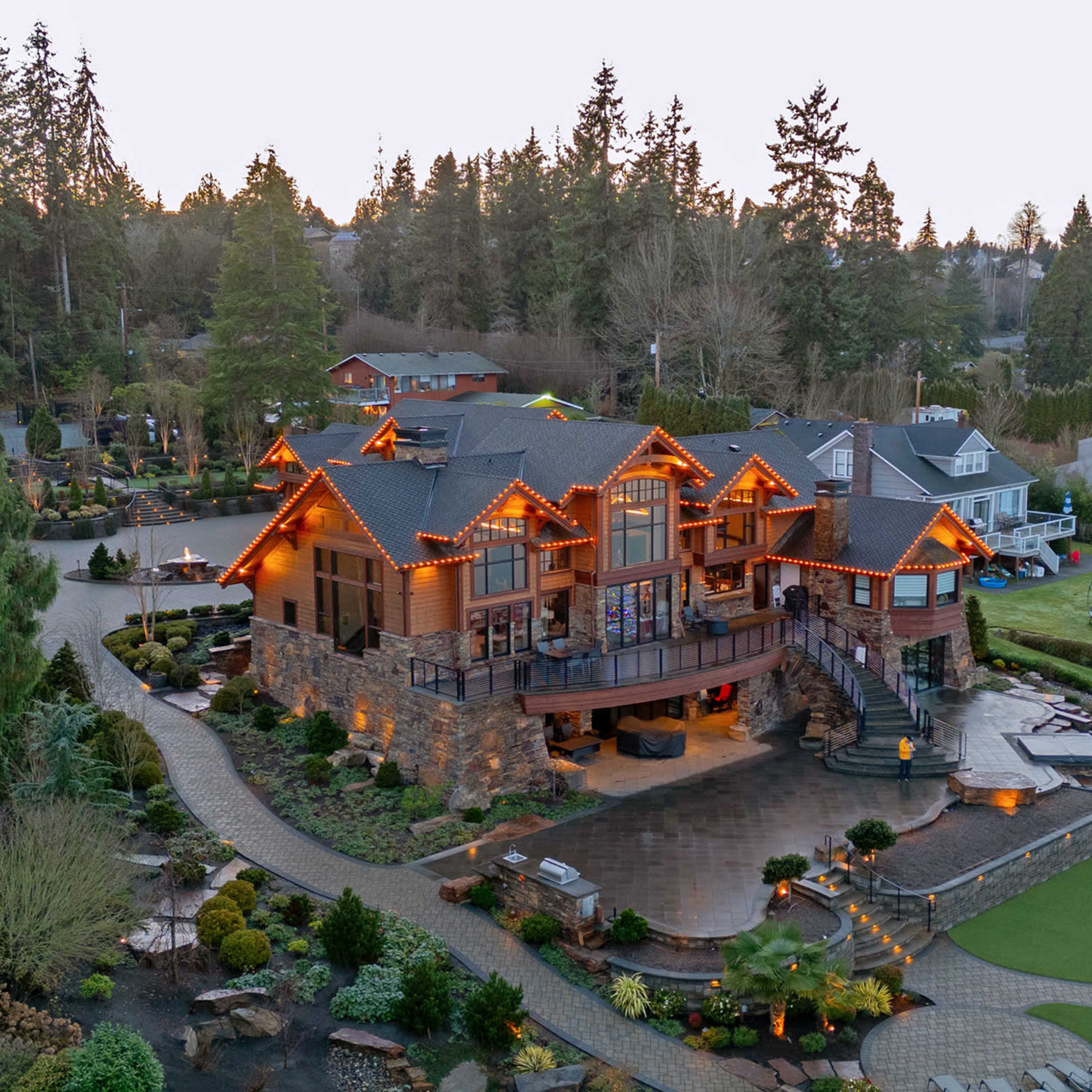 A large, multi-level house decorated with orange lights is surrounded by well-maintained gardens and trees.