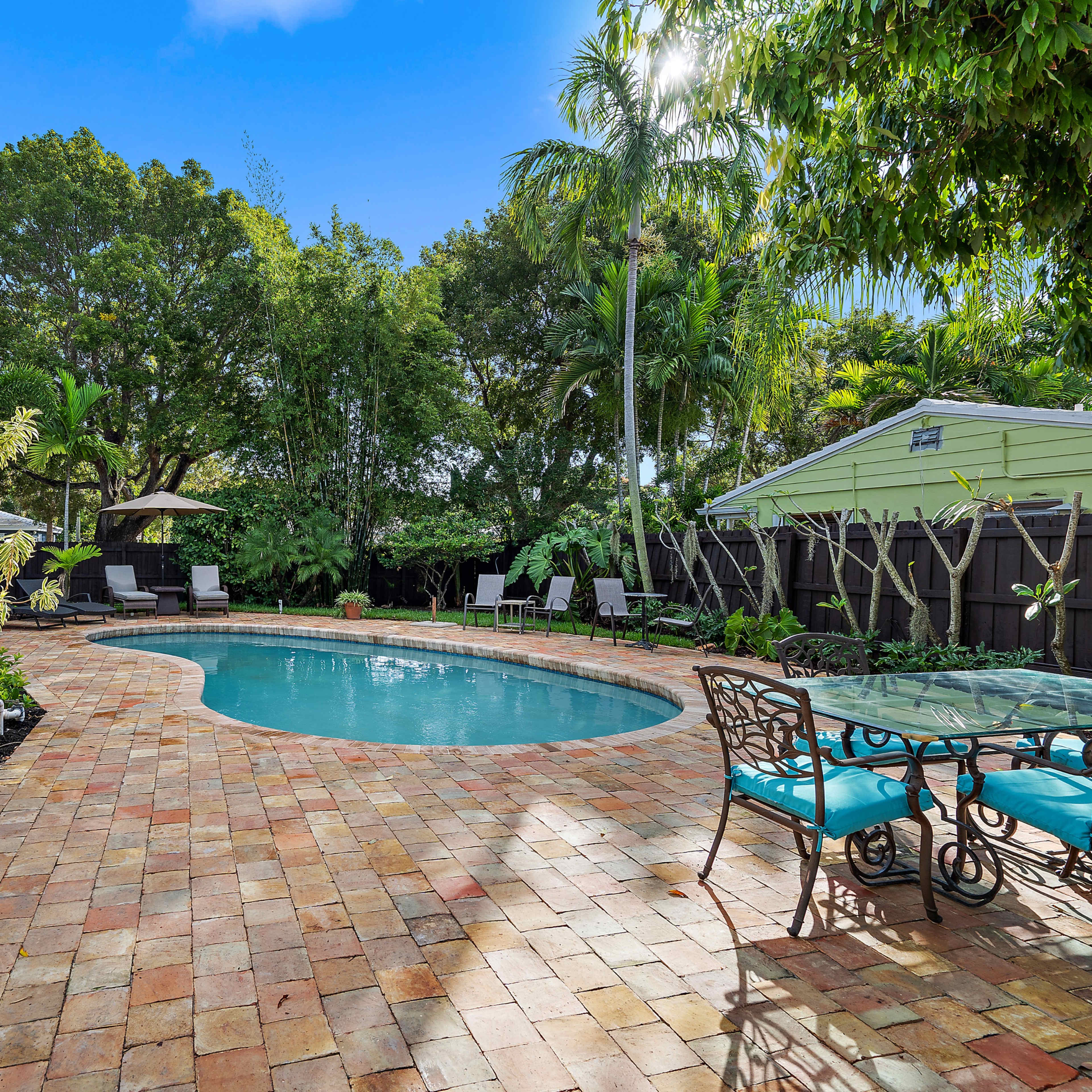 A backyard with a swimming pool surrounded by a tiled patio and lounge chairs, framed by lush greenery and shaded by trees.