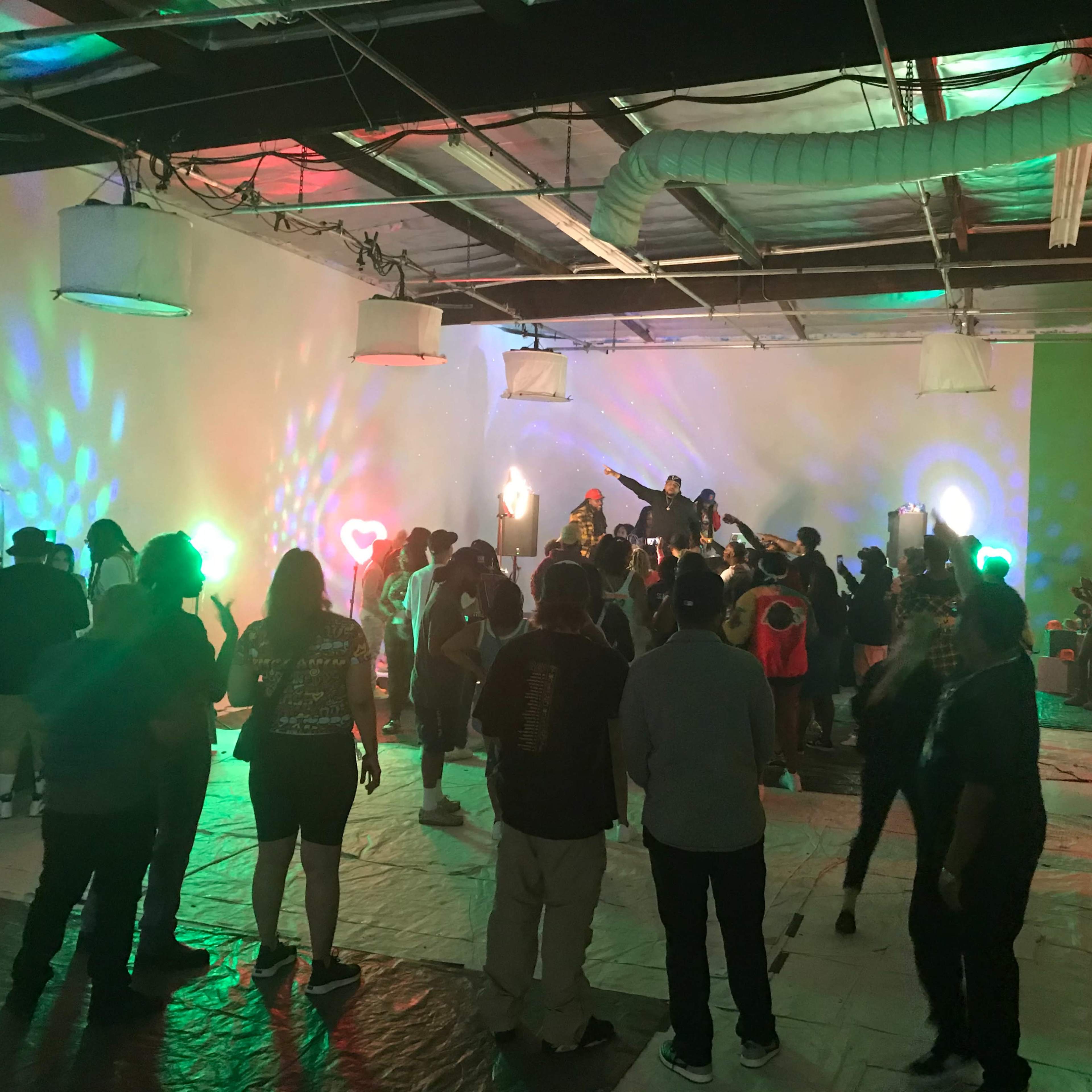 A crowd gathers in a brightly lit indoor space with colorful lights and a stage featuring performers.