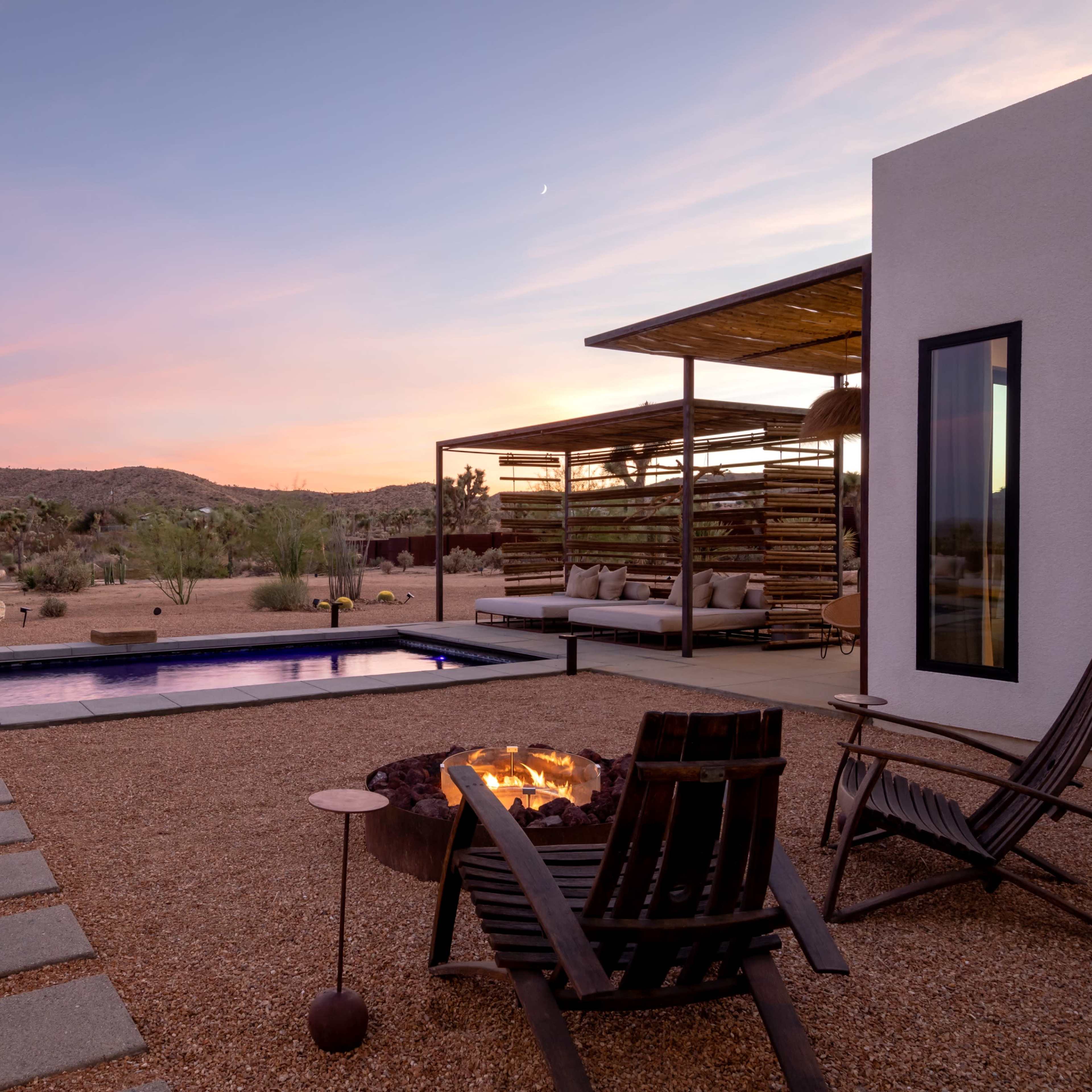 A modern outdoor space features a swimming pool, fire pit, and seating area set against a desert landscape at sunset.