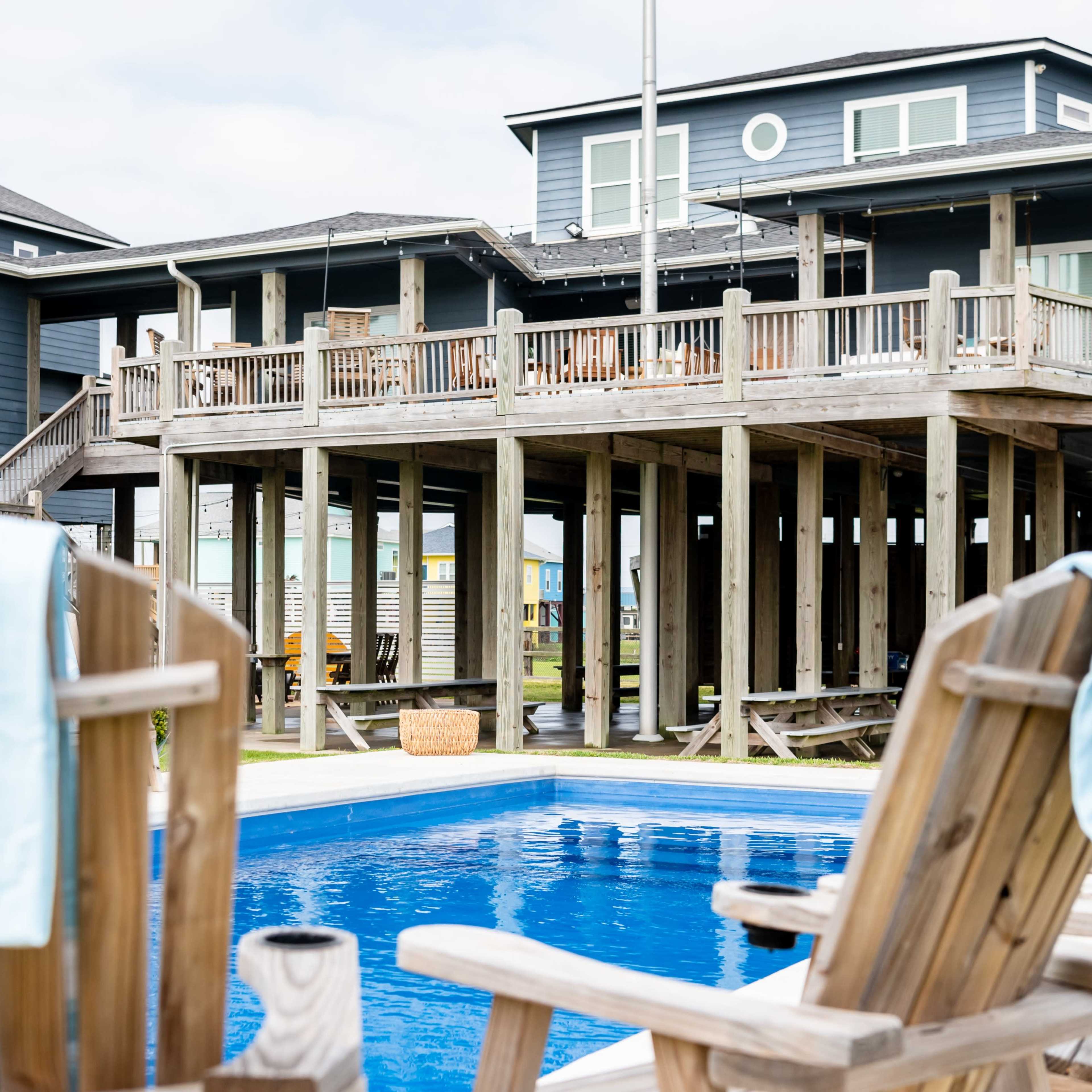 A wooden deck house elevated on stilts overlooks a swimming pool with two lounge chairs in the foreground.