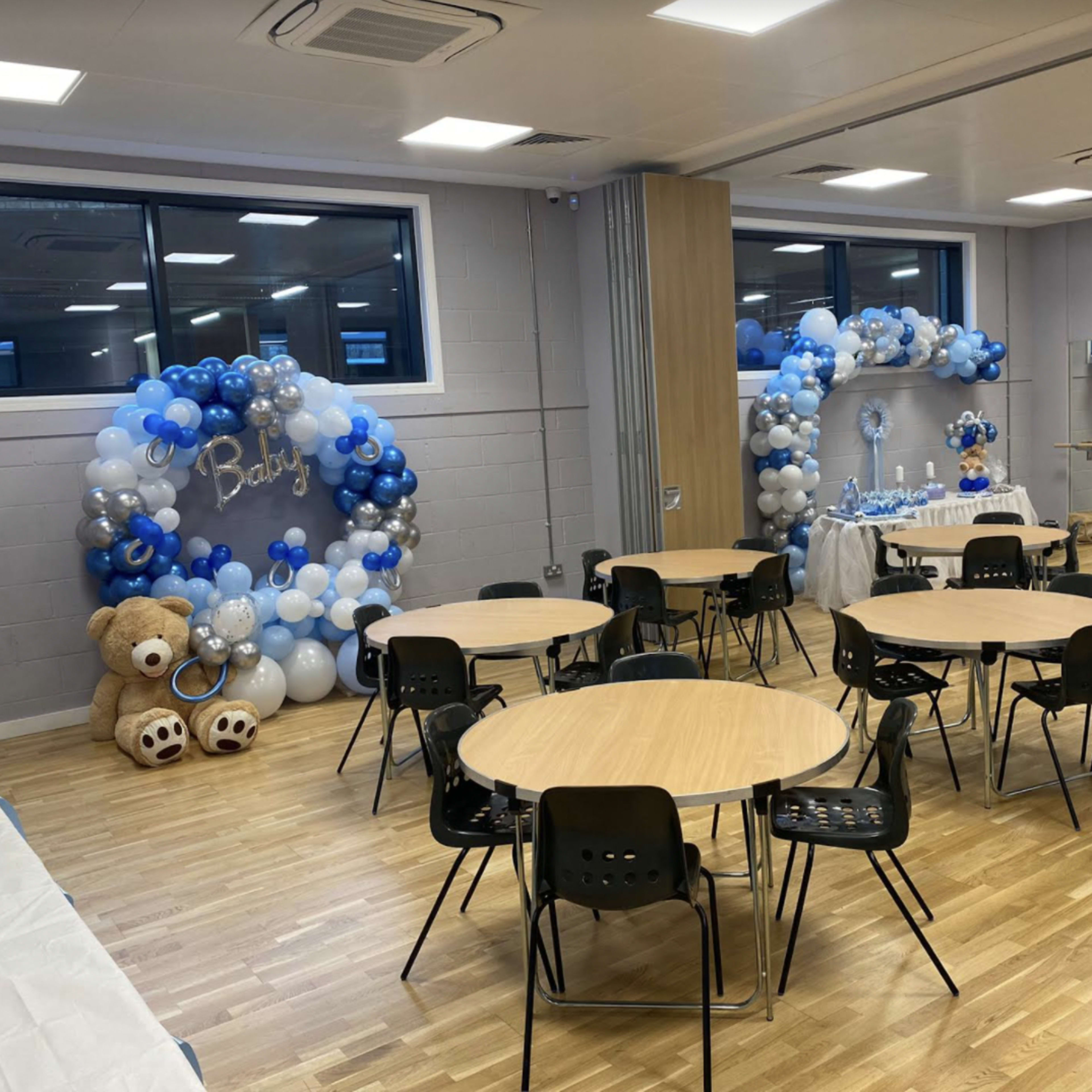 A decorated event space featuring balloon arches in shades of blue, tables with black chairs, and teddy bear centerpieces.