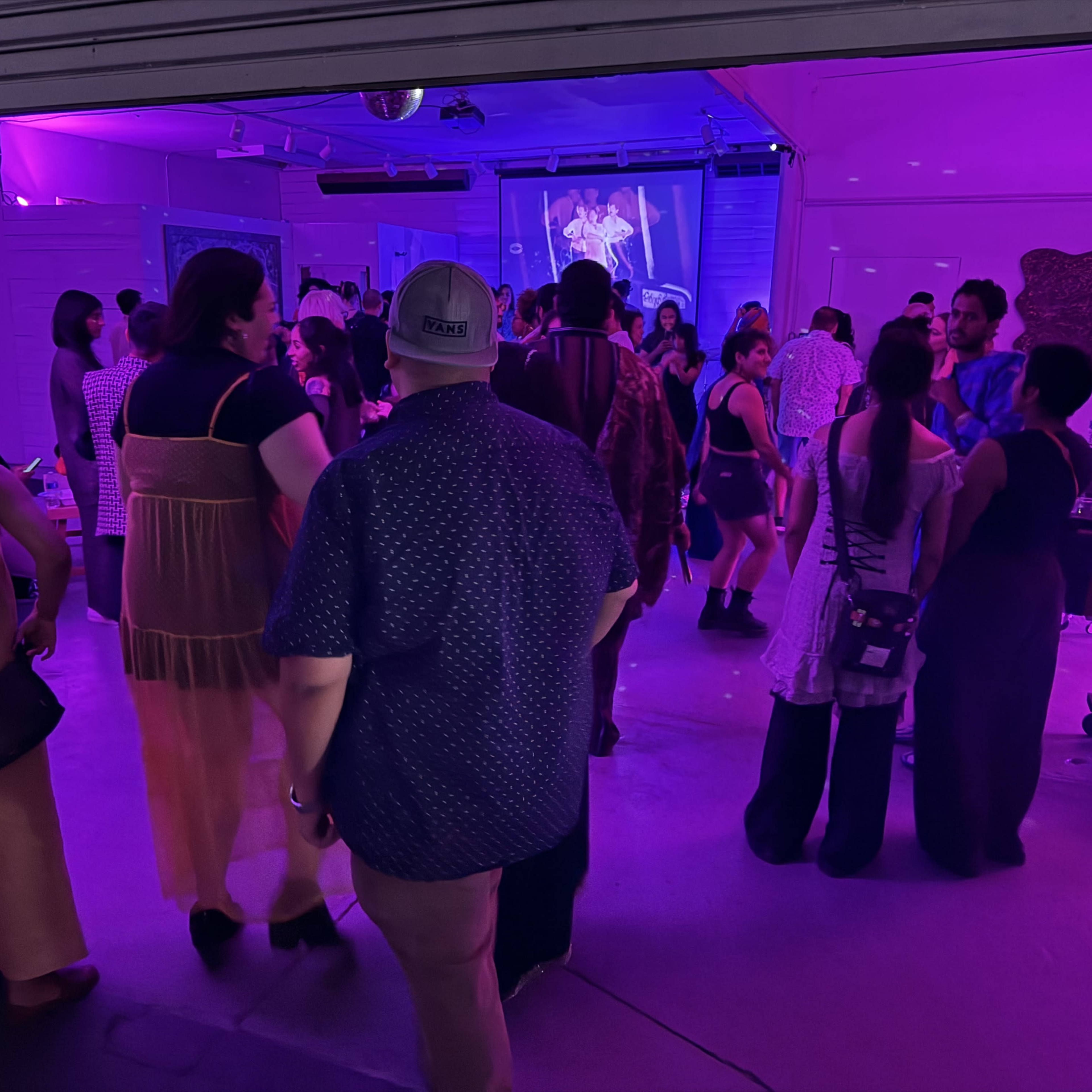 A crowd of people mingles in a dimly lit space illuminated by purple lights, with a projection on a wall in the background.