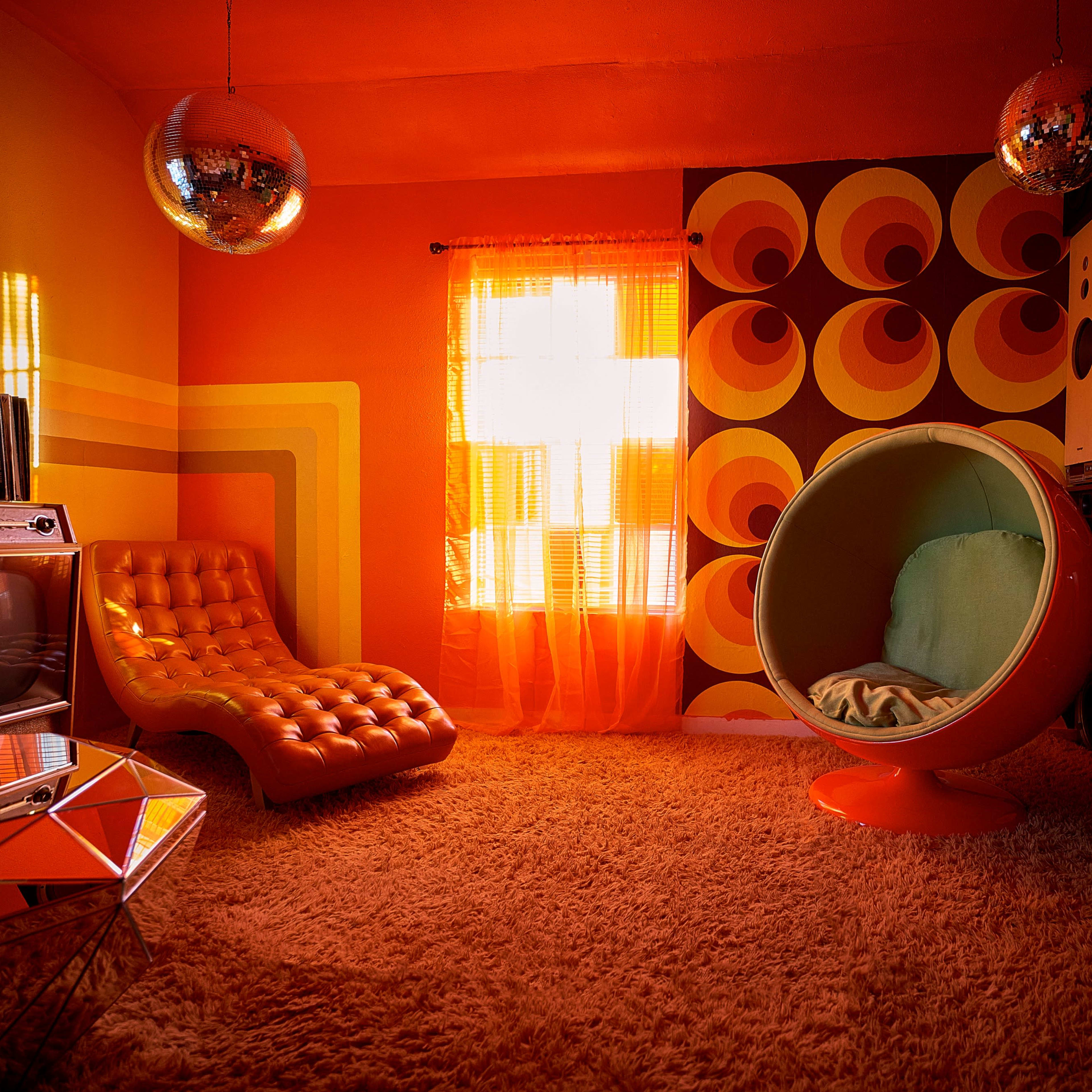 A retro-styled room with orange walls, a circular chair, a vintage television set, and large speakers, all complemented by disco balls and a shag carpet.