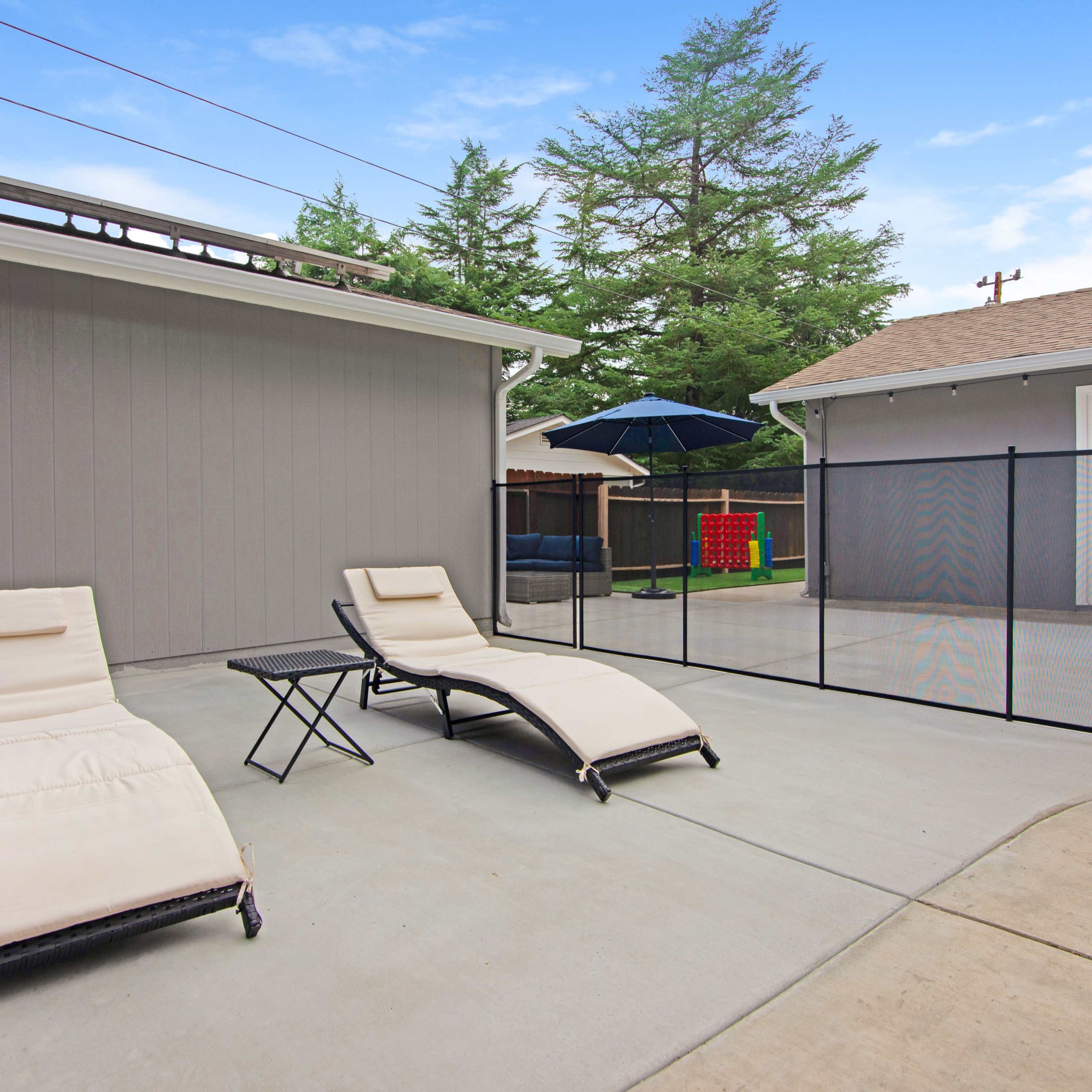 A fenced outdoor patio area with two lounge chairs, a small table, and a large umbrella beside two houses.