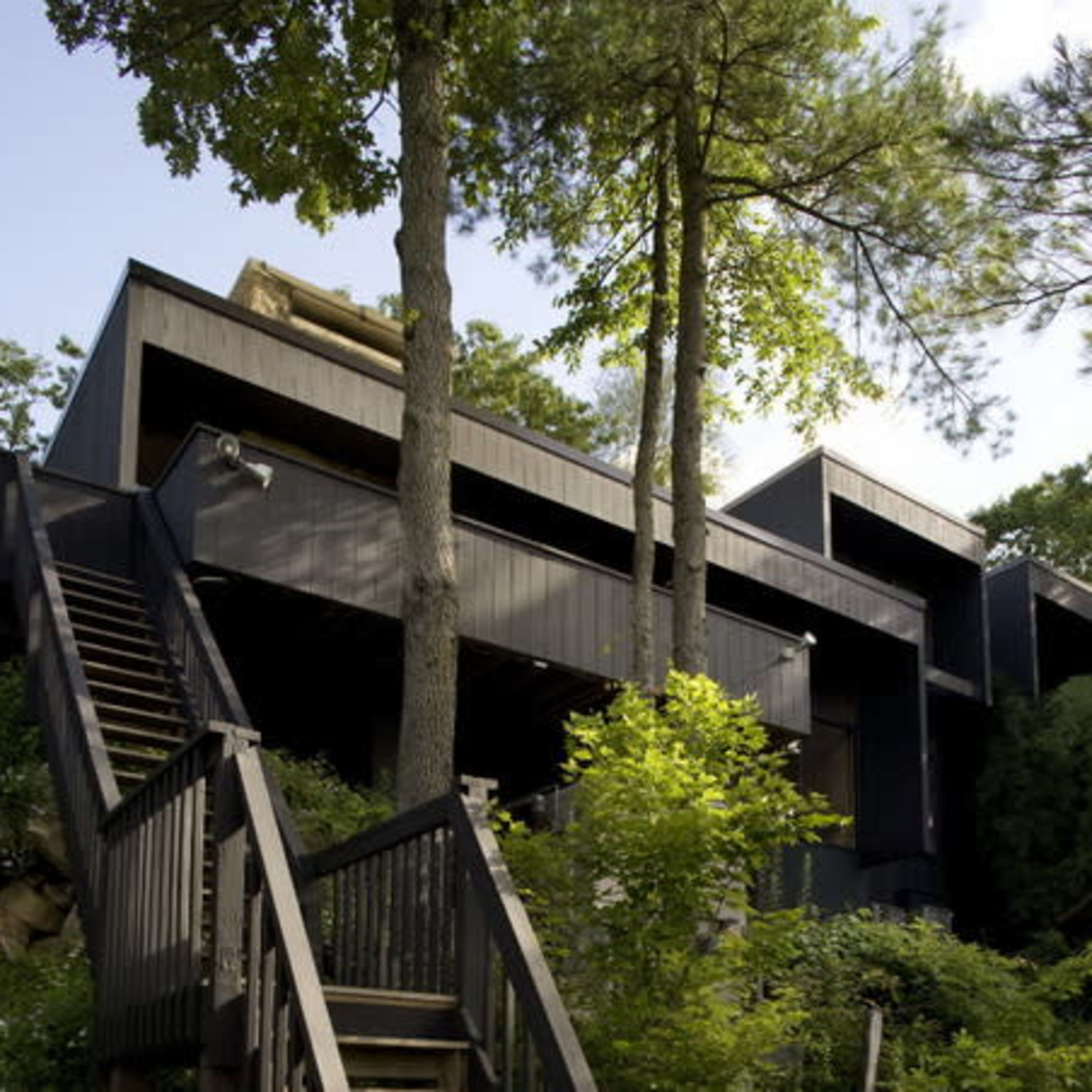 A modern, dark-colored house built on a hillside, surrounded by trees and greenery, with a wooden staircase leading up to it.