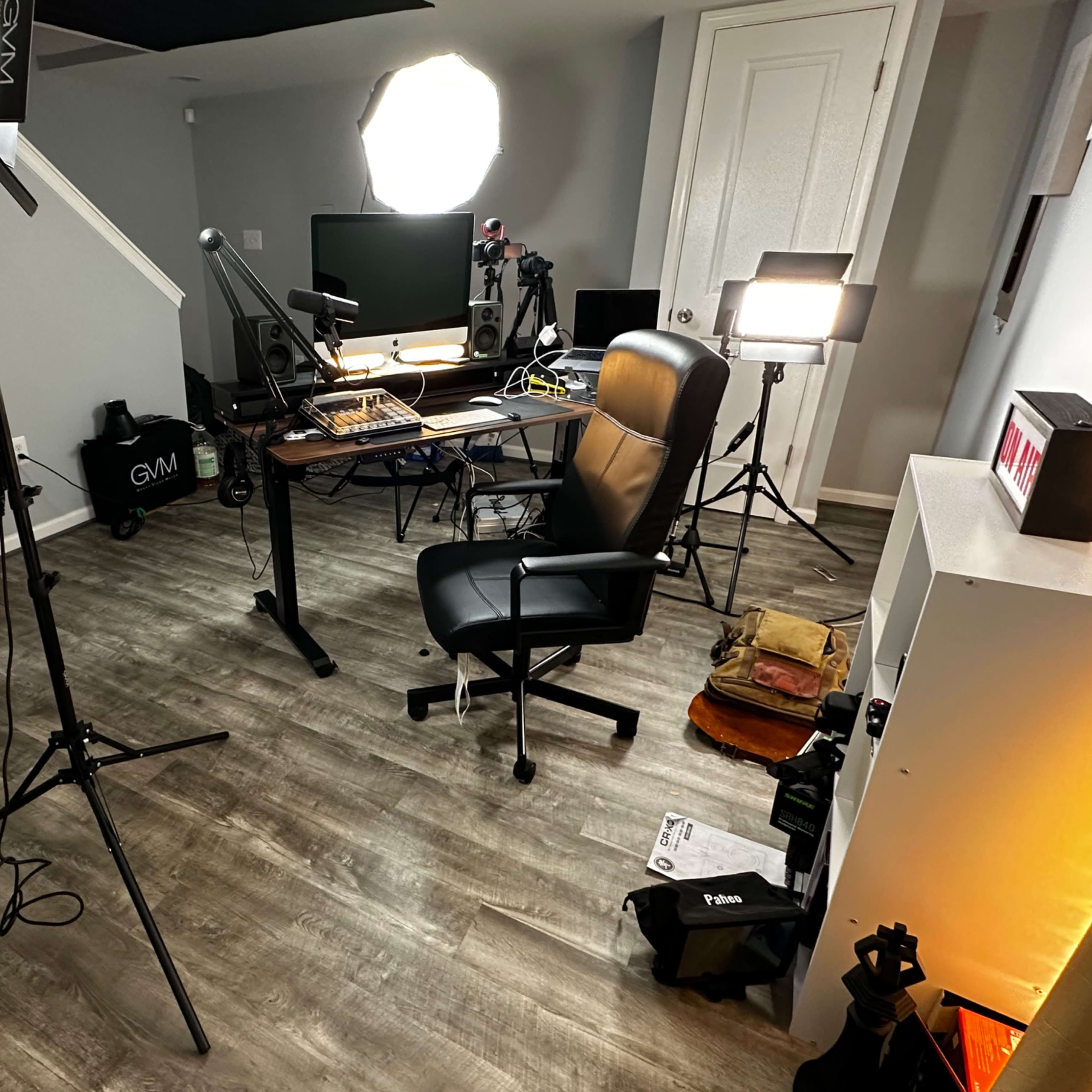 A cluttered home office setup features a desk with computer equipment, lighting stands, and scattered items on the floor.