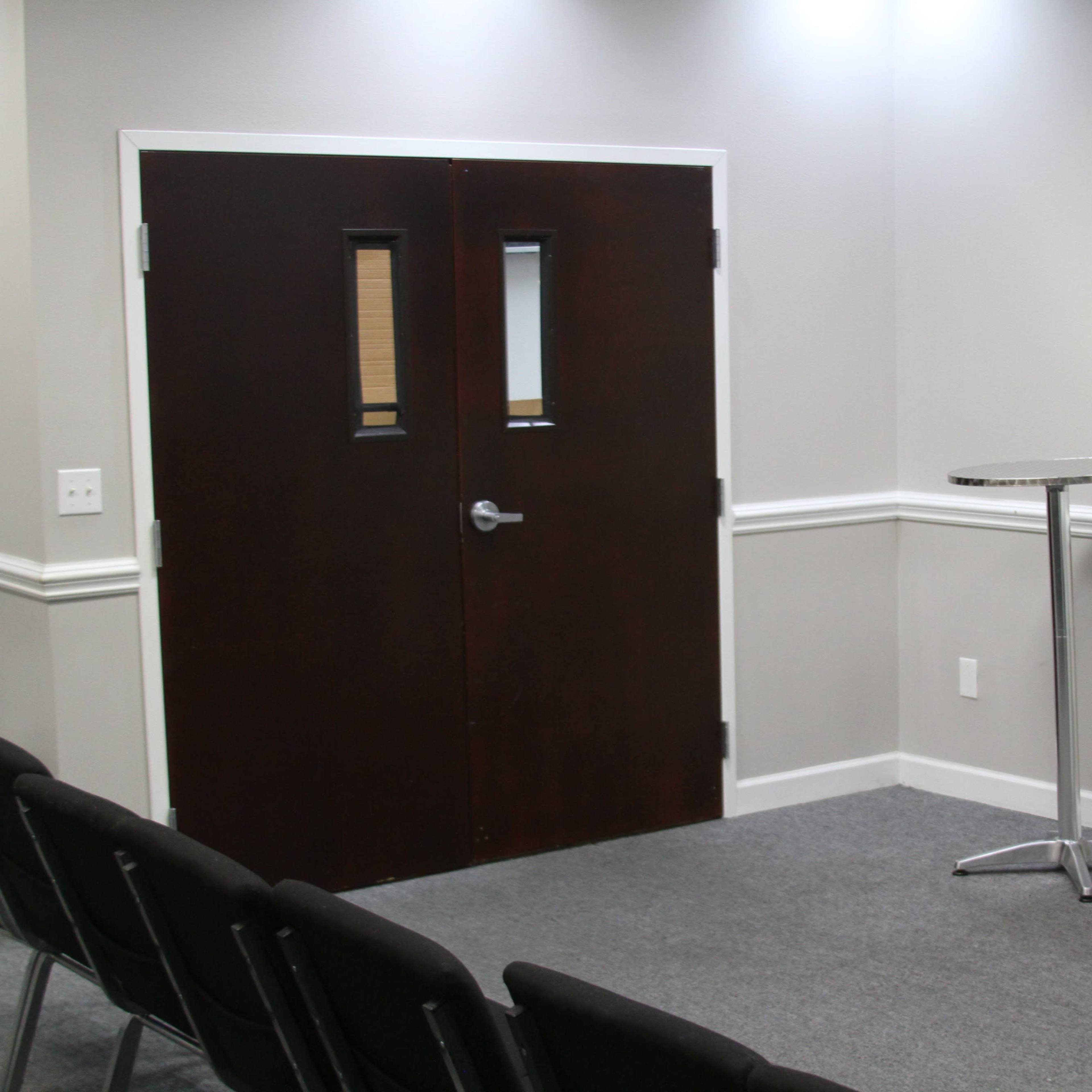 A small meeting room featuring a dark wooden door, a podium, a standing fan, and a television mounted on the wall, with rows of chairs arranged in front.