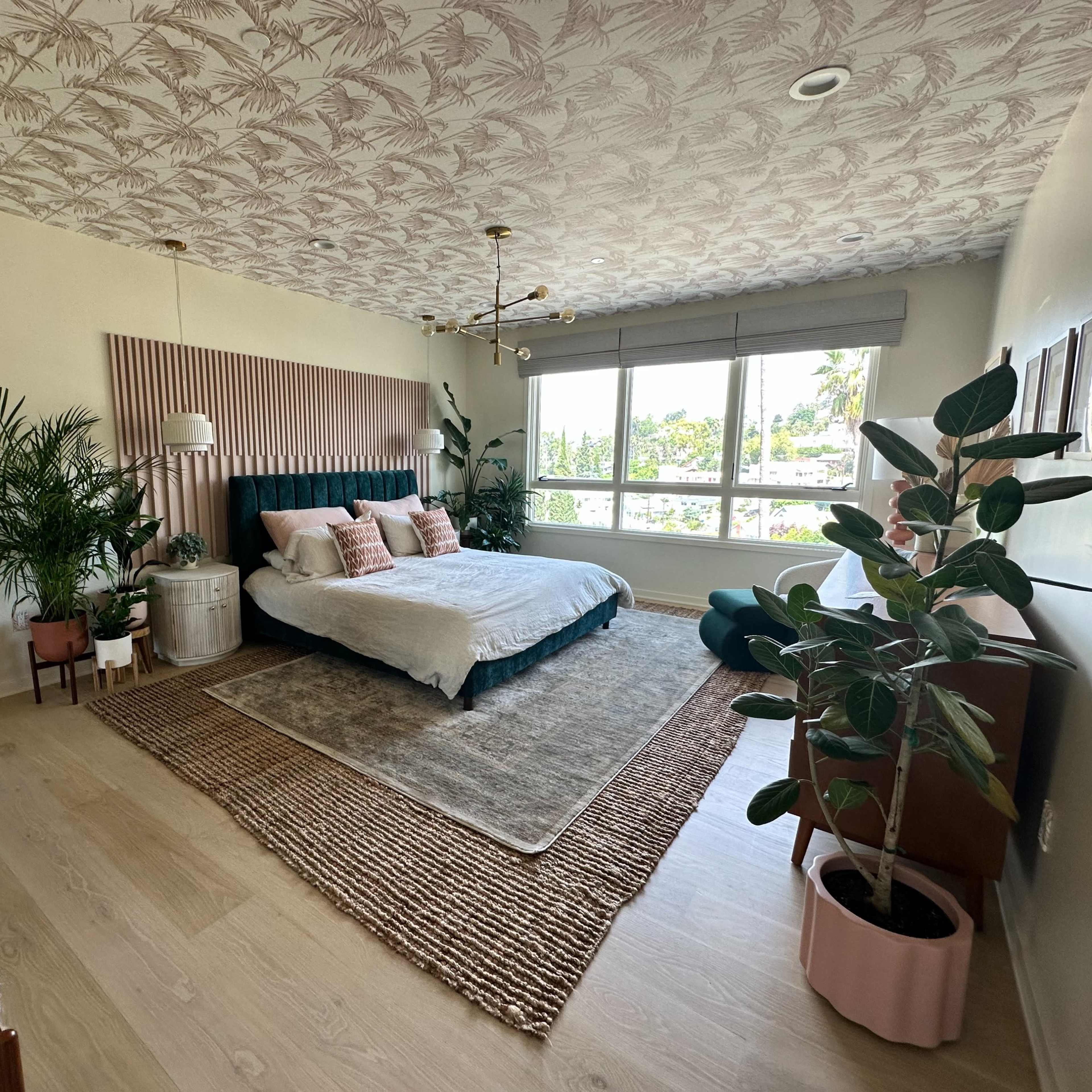 A spacious bedroom features a large bed with decorative pillows, plants, and a light rug, framed by windows that allow natural light to enter.