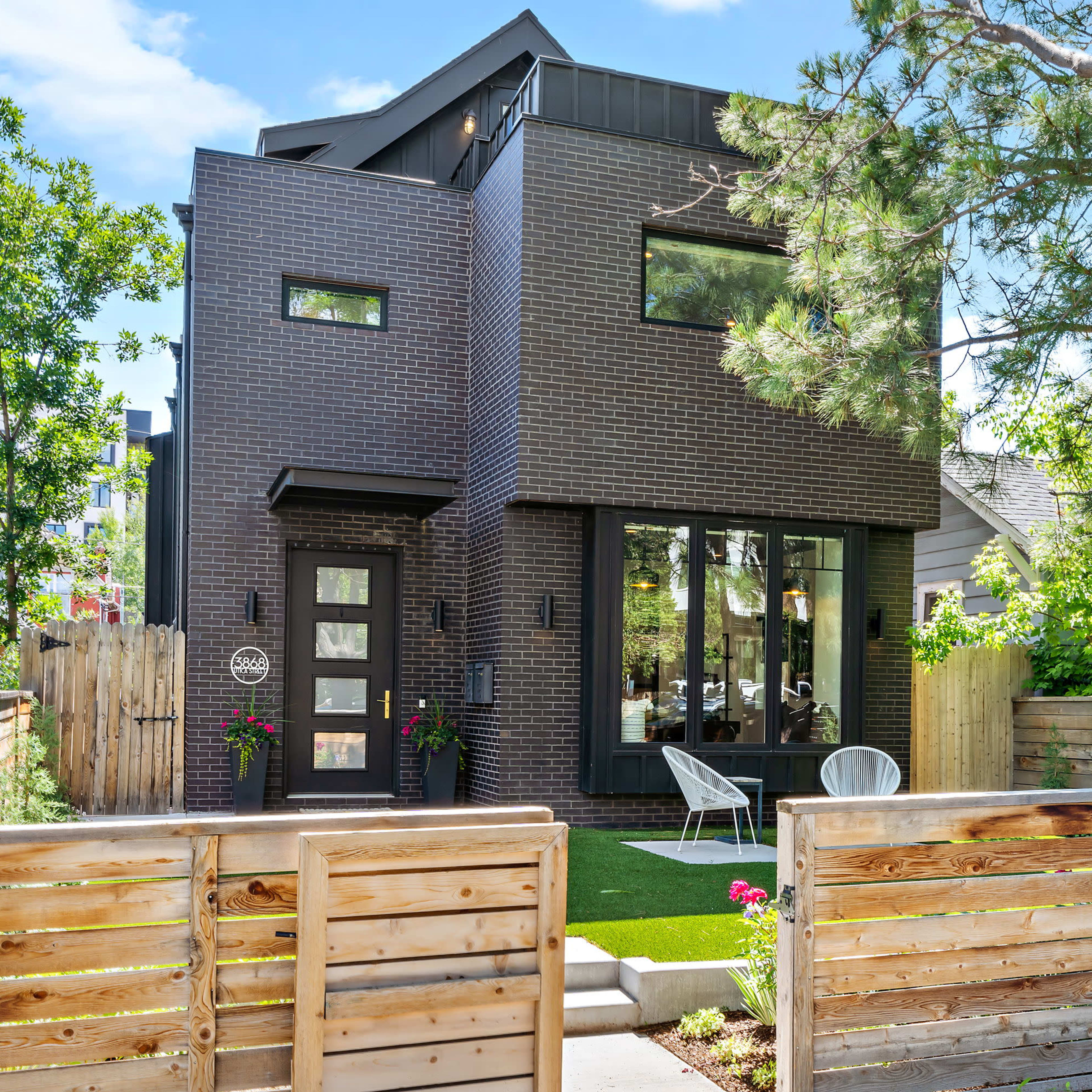 A modern two-story house with dark brick exterior, large windows, and a landscaped front yard enclosed by a wooden fence.