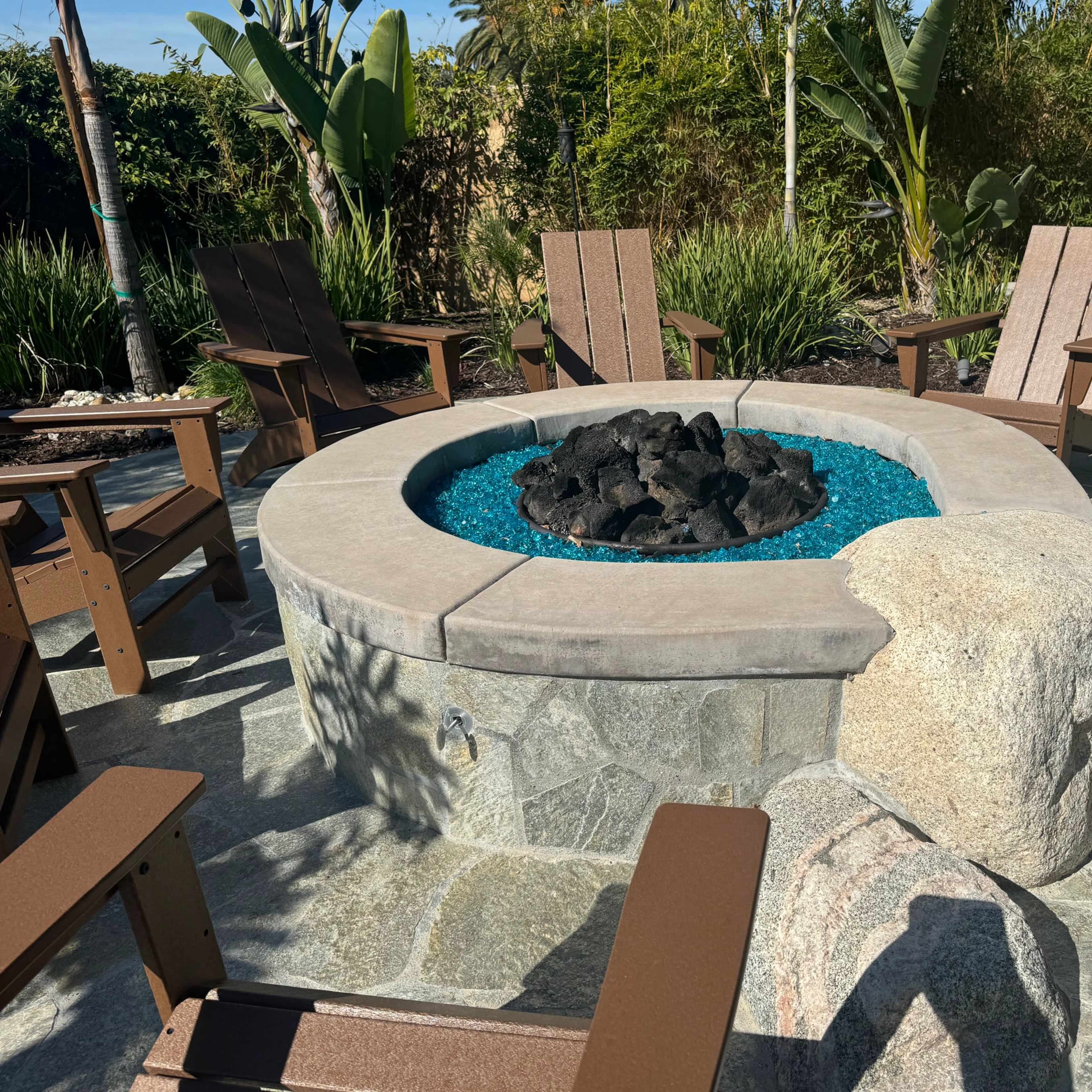 A circular fire pit surrounded by wooden chairs and blue glass stones, set in a landscaped outdoor area with plants.