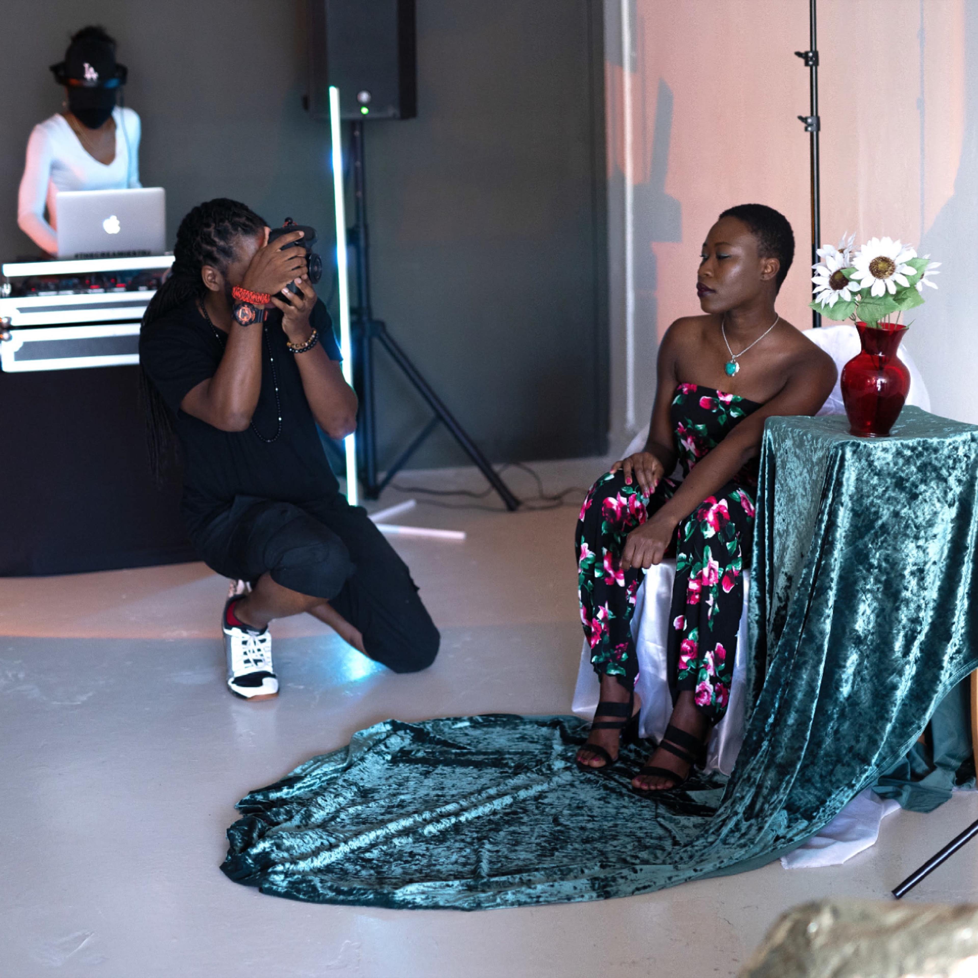 A photographer kneels to take a picture of a woman seated on a table draped in green fabric, while a DJ sets up in the background.