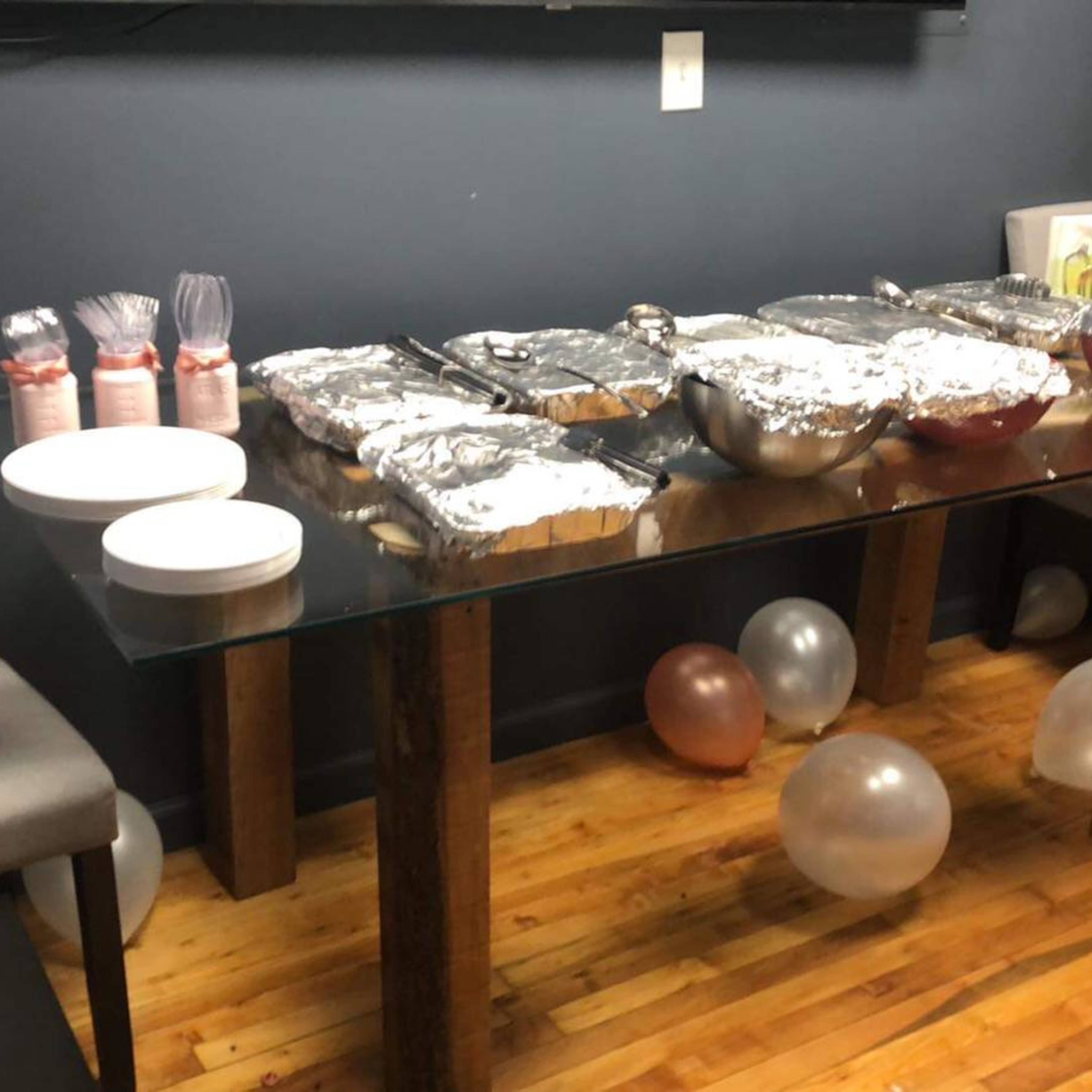 A table is set with covered food containers, drinks, plates, and balloons in a modest room.