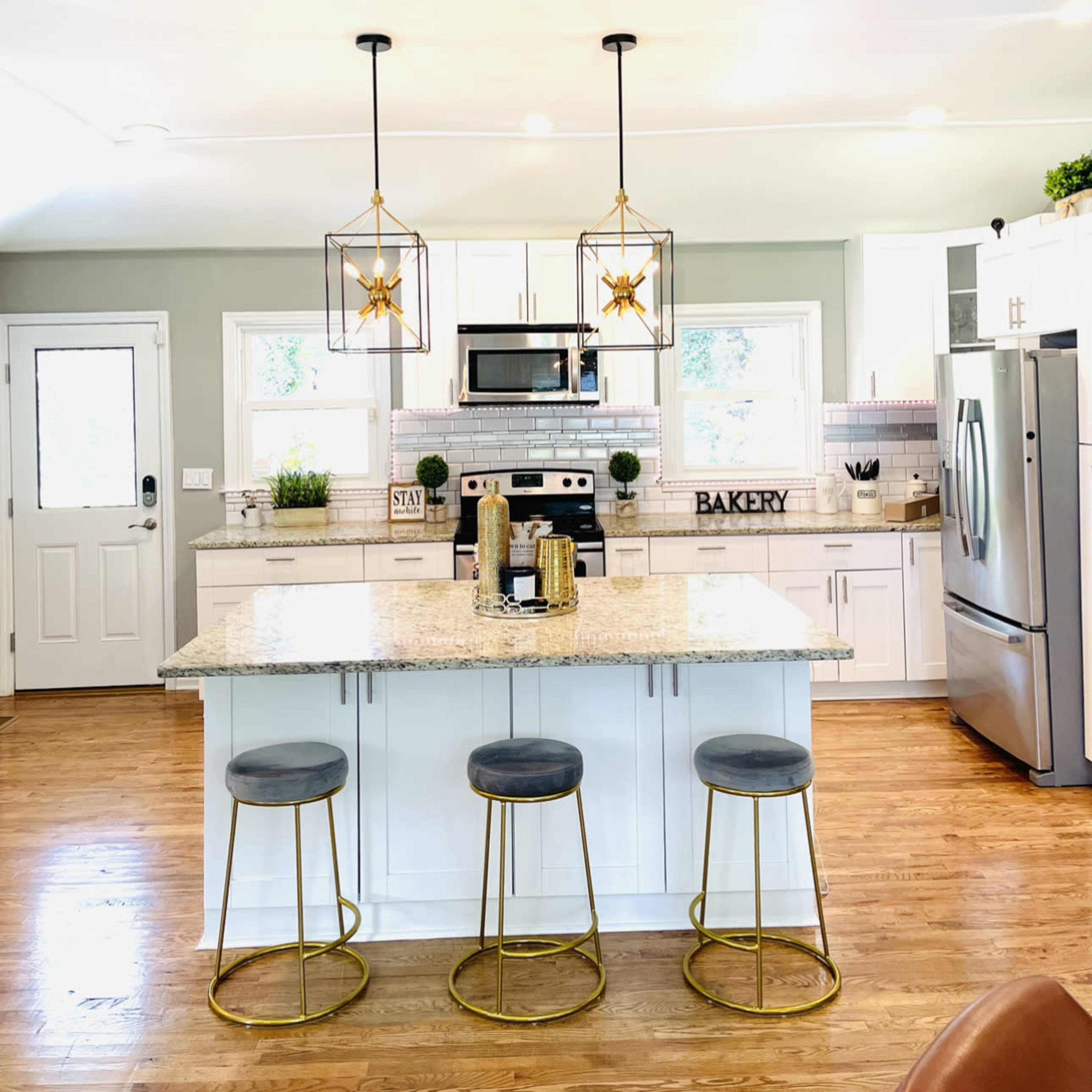 A modern kitchen features a large island with three gold-accented stools and two decorative pendant lights above.
