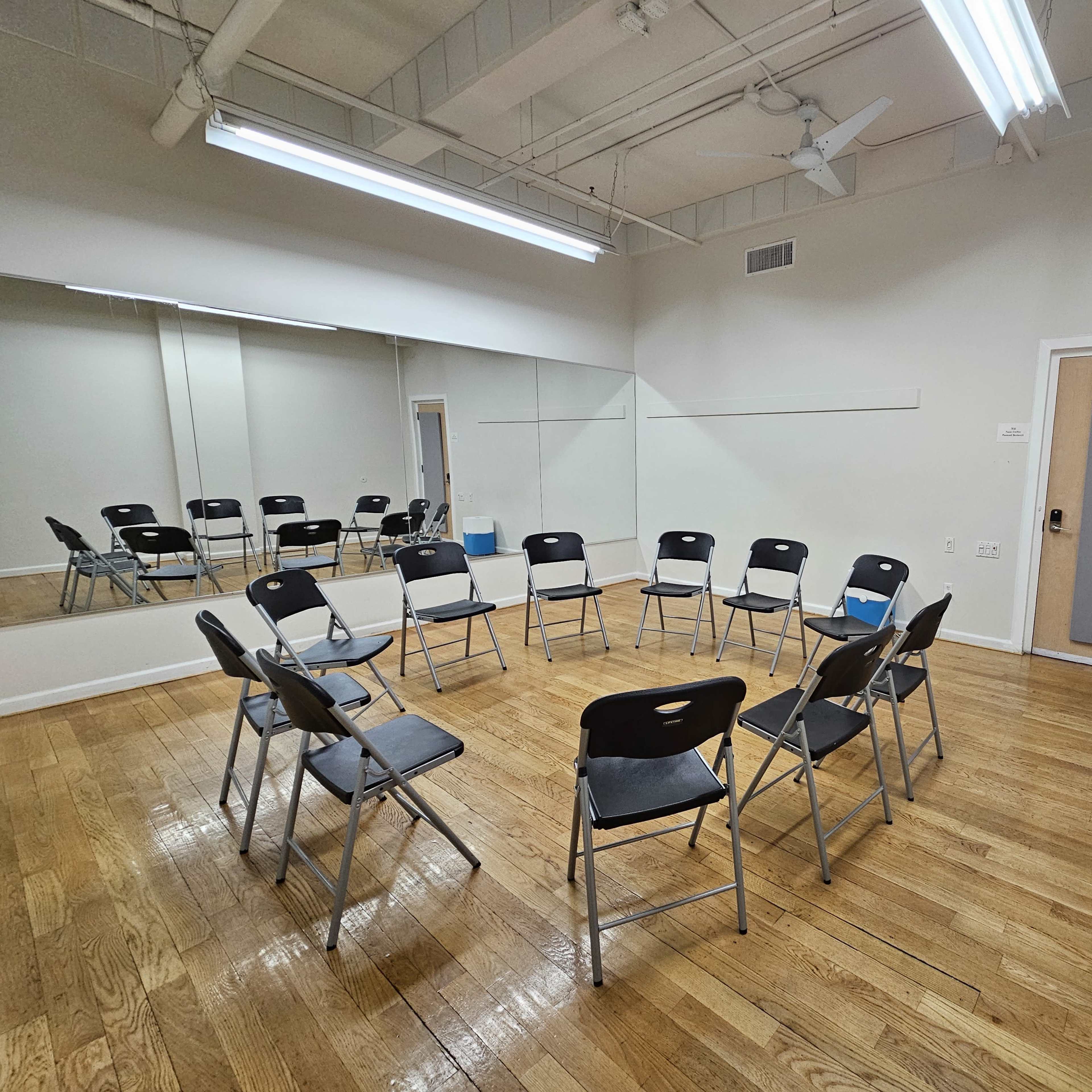 A room with wooden flooring, featuring a circle of black folding chairs and a large mirror on one wall.
