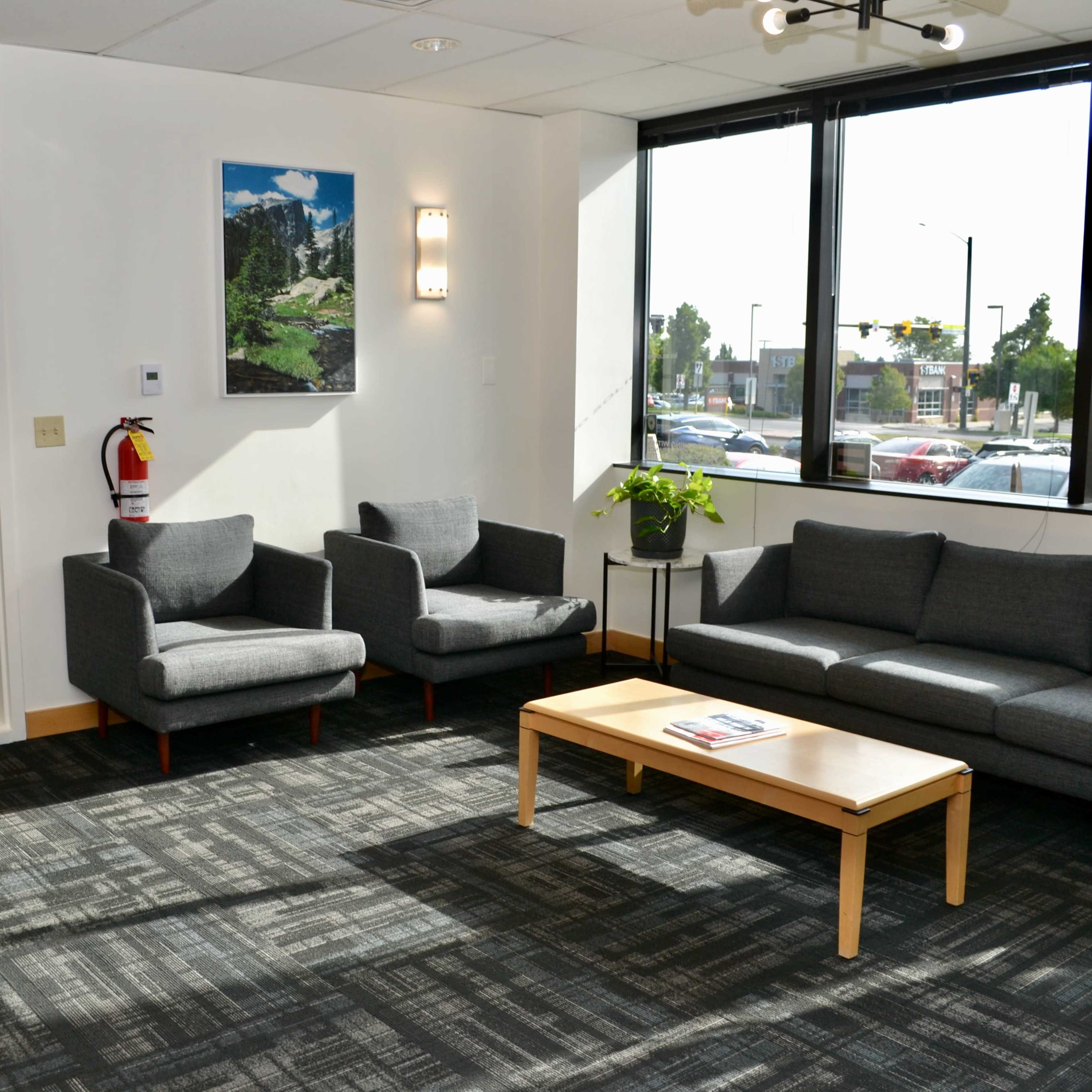 A modern waiting area with two armchairs, a sofa, and a coffee table under large windows overlooking a street.