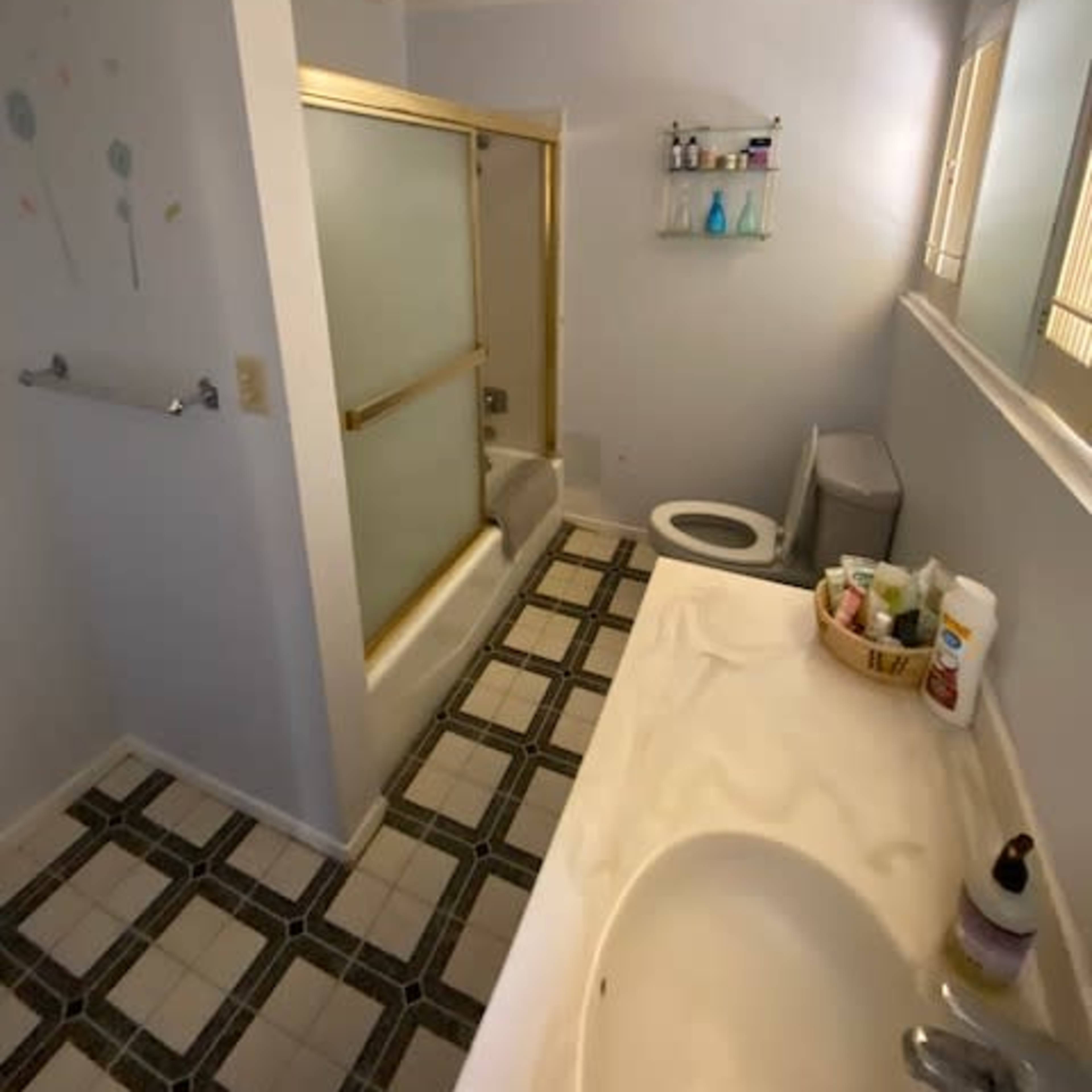 A small bathroom featuring a bathtub with a shower enclosure, a toilet, a sink with various toiletries, and a window allowing natural light.
