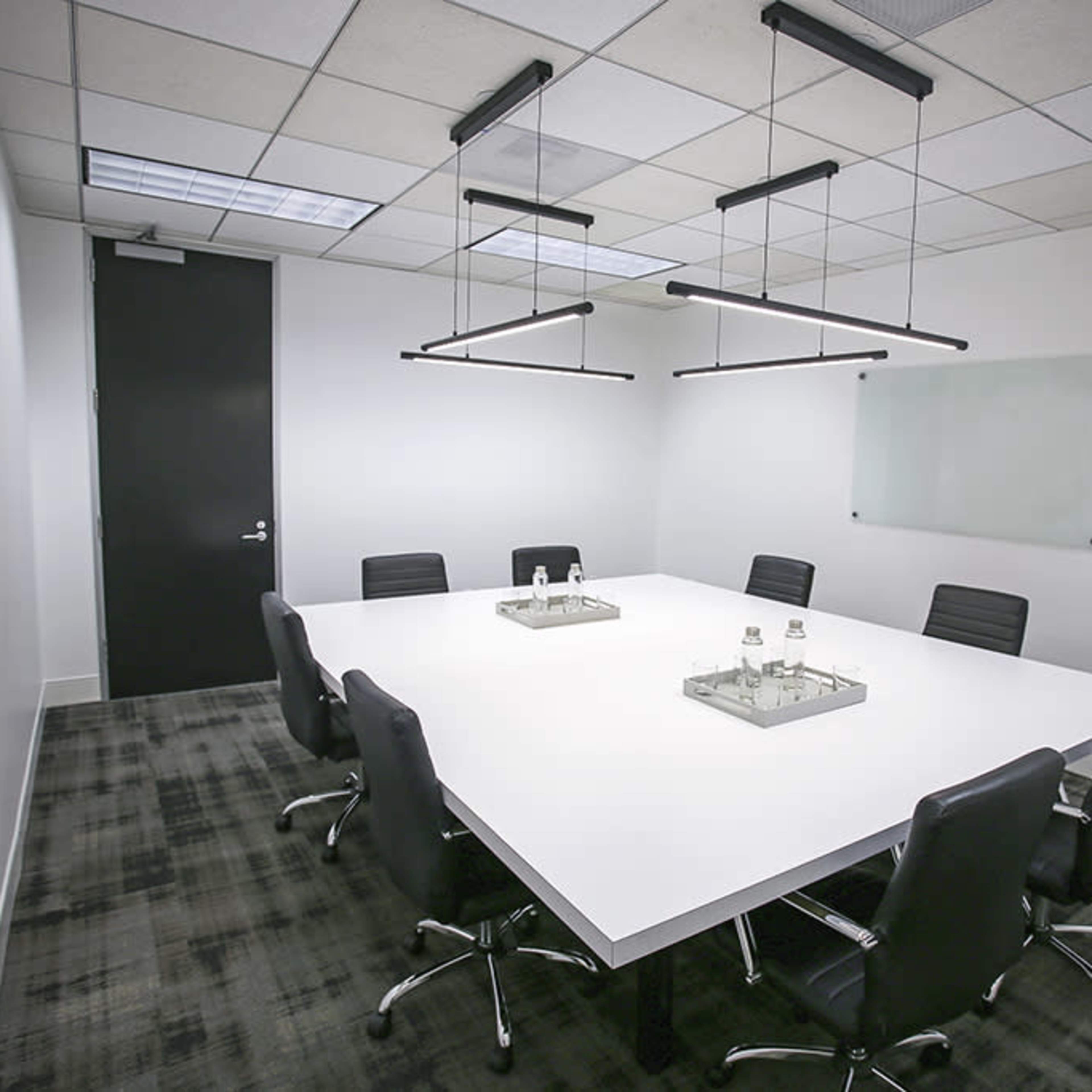 A modern conference room features a large white table surrounded by black chairs, with minimalistic hanging lights and a glass board on the wall.