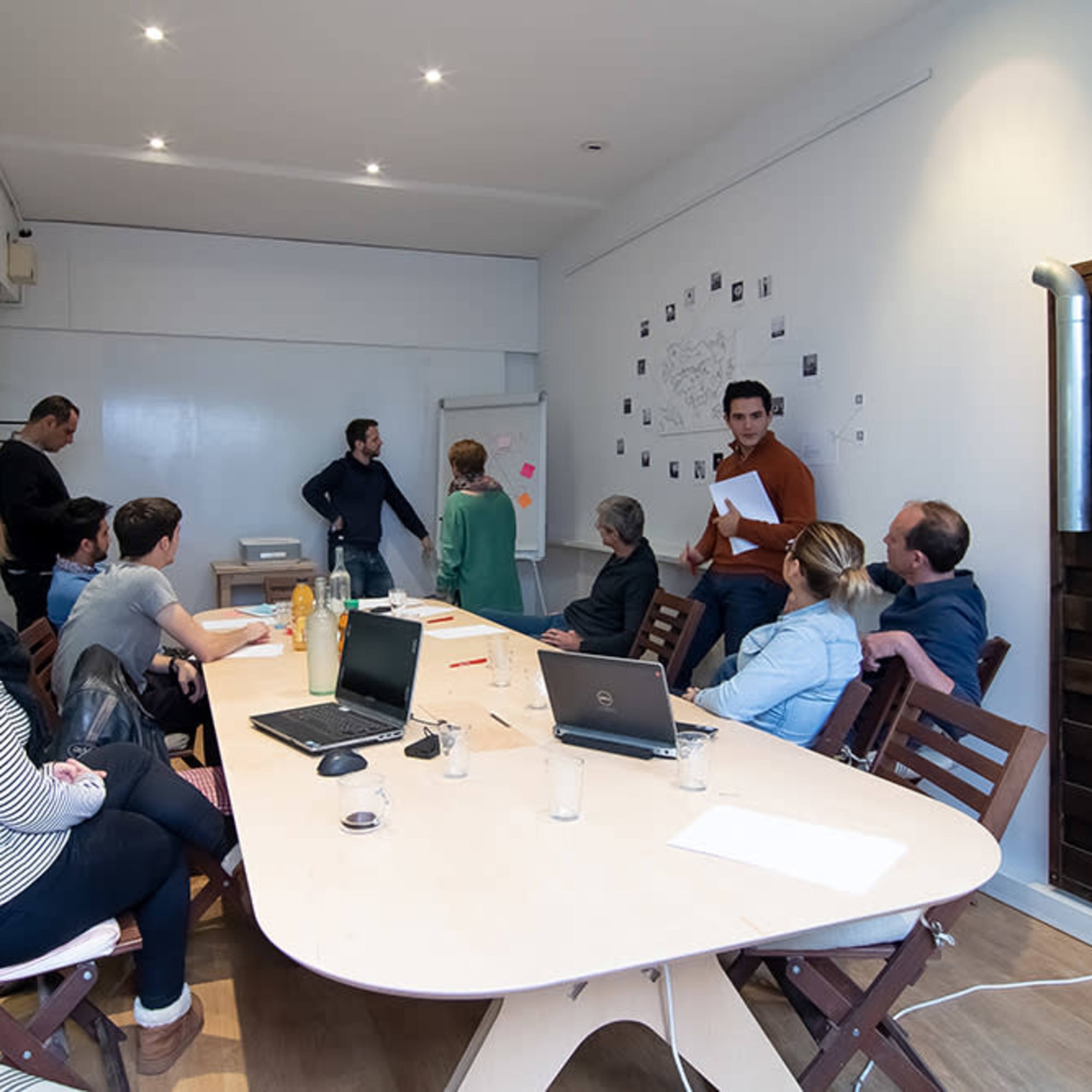 A group of ten people engages in a discussion around a large table in a meeting room with a whiteboard and wall-mounted plants.
