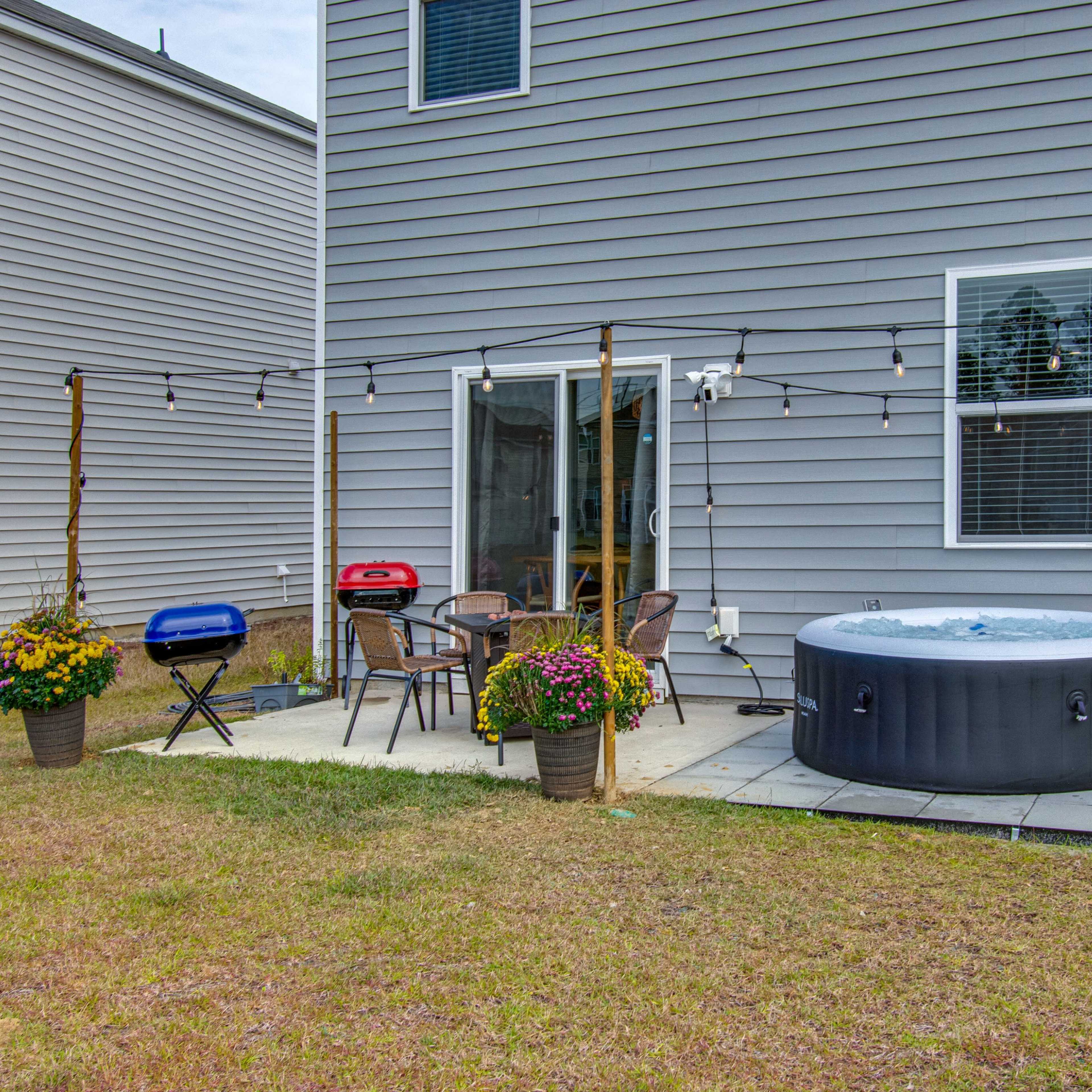 A patio area with a hot tub, grill, and dining chairs surrounded by potted plants in front of a house with gray siding.