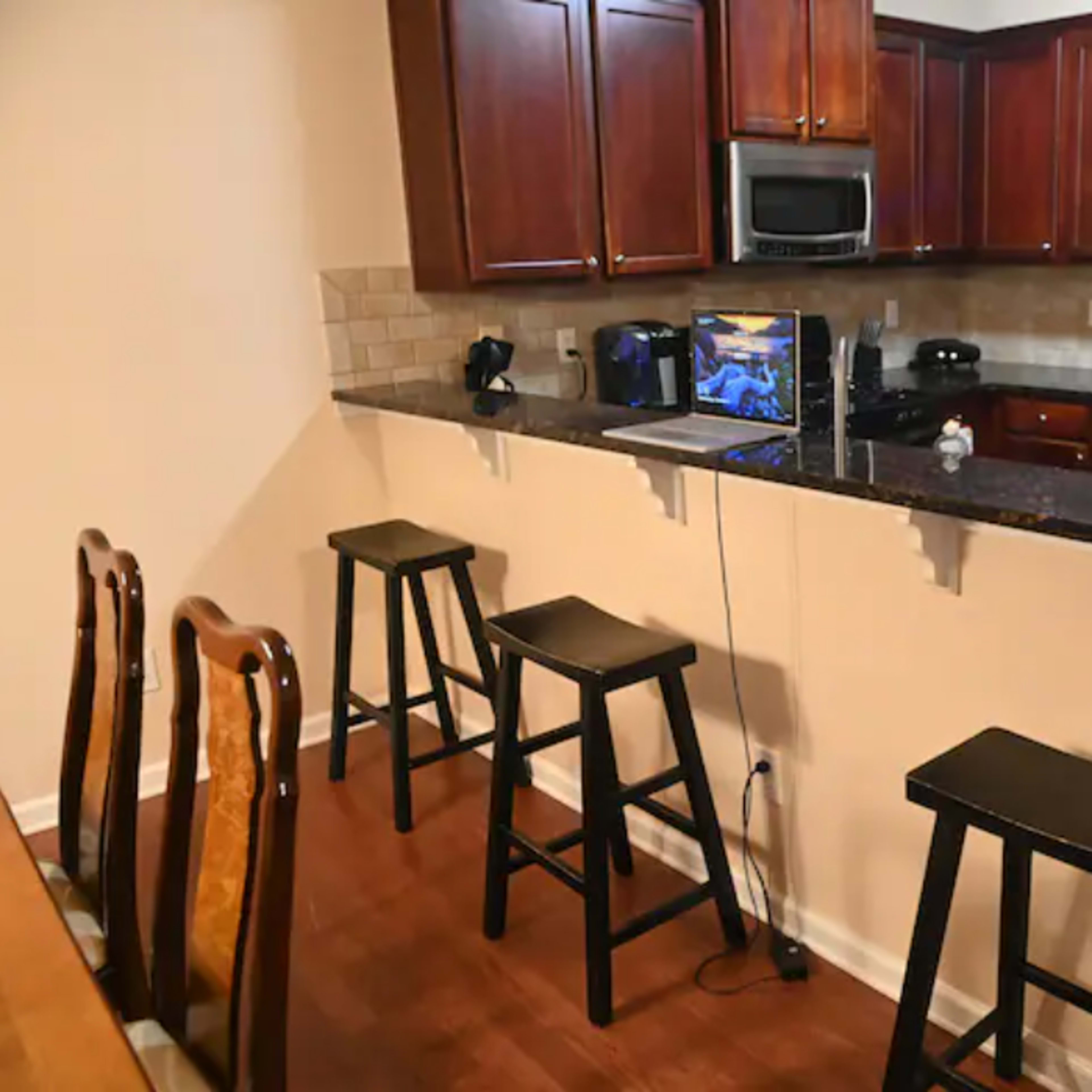 A modern kitchen features a granite countertop with four black stools, an open laptop, and stainless steel appliances under wooden cabinets.