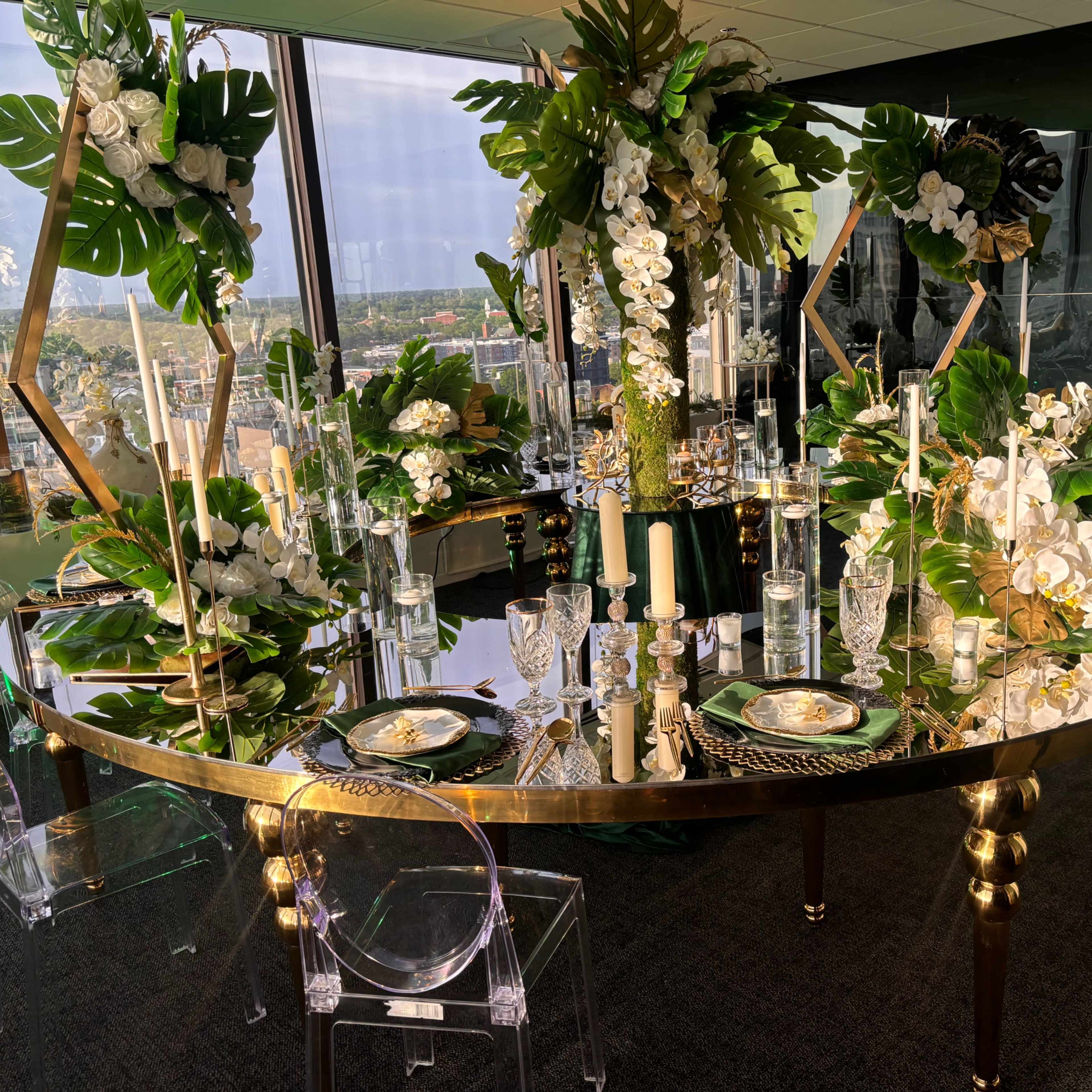A round dining table is elegantly set with floral arrangements, candles, and glassware against a panoramic city view.