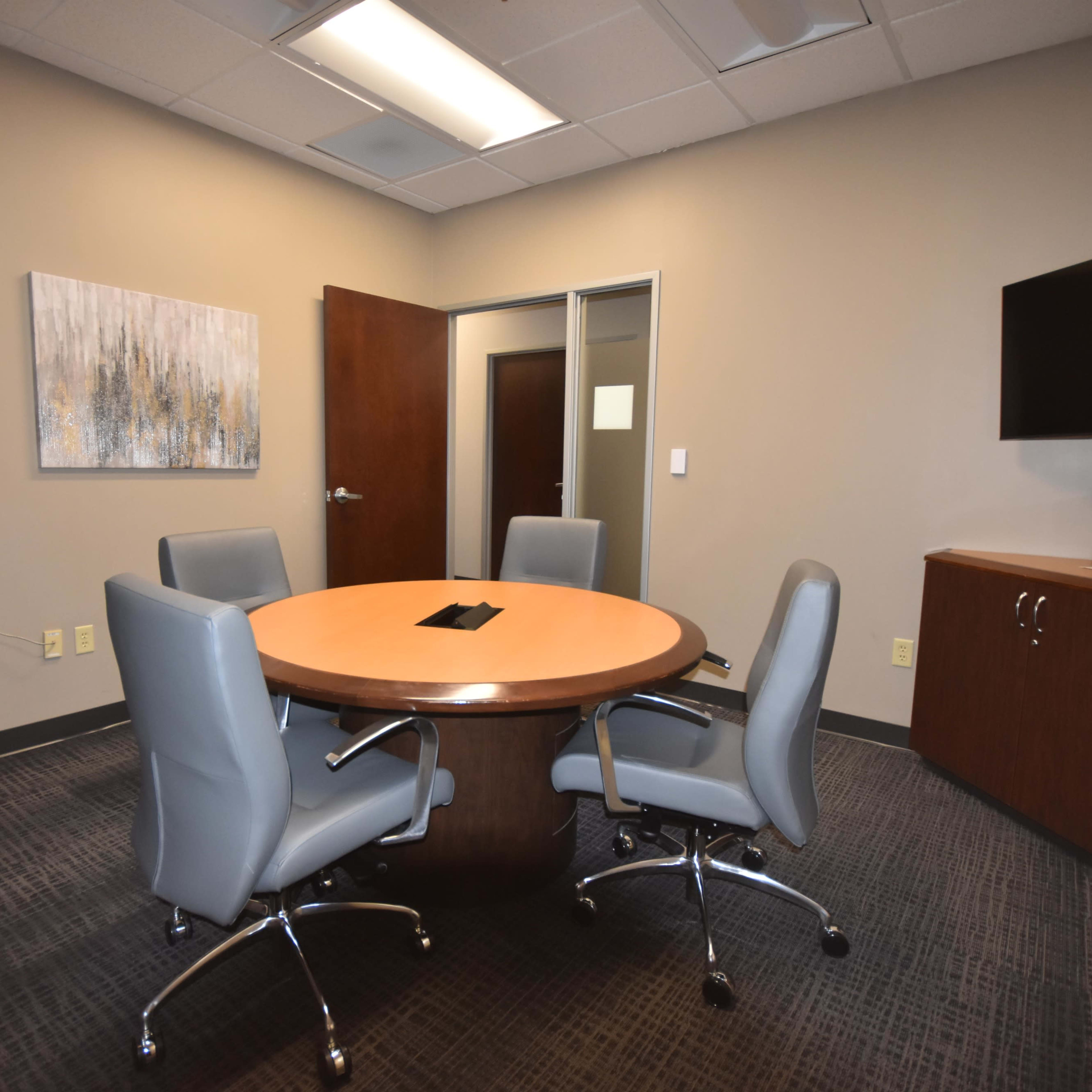 A corporate meeting room featuring a circular wooden table surrounded by four gray chairs, with a television mounted on the wall and cabinets on either side.