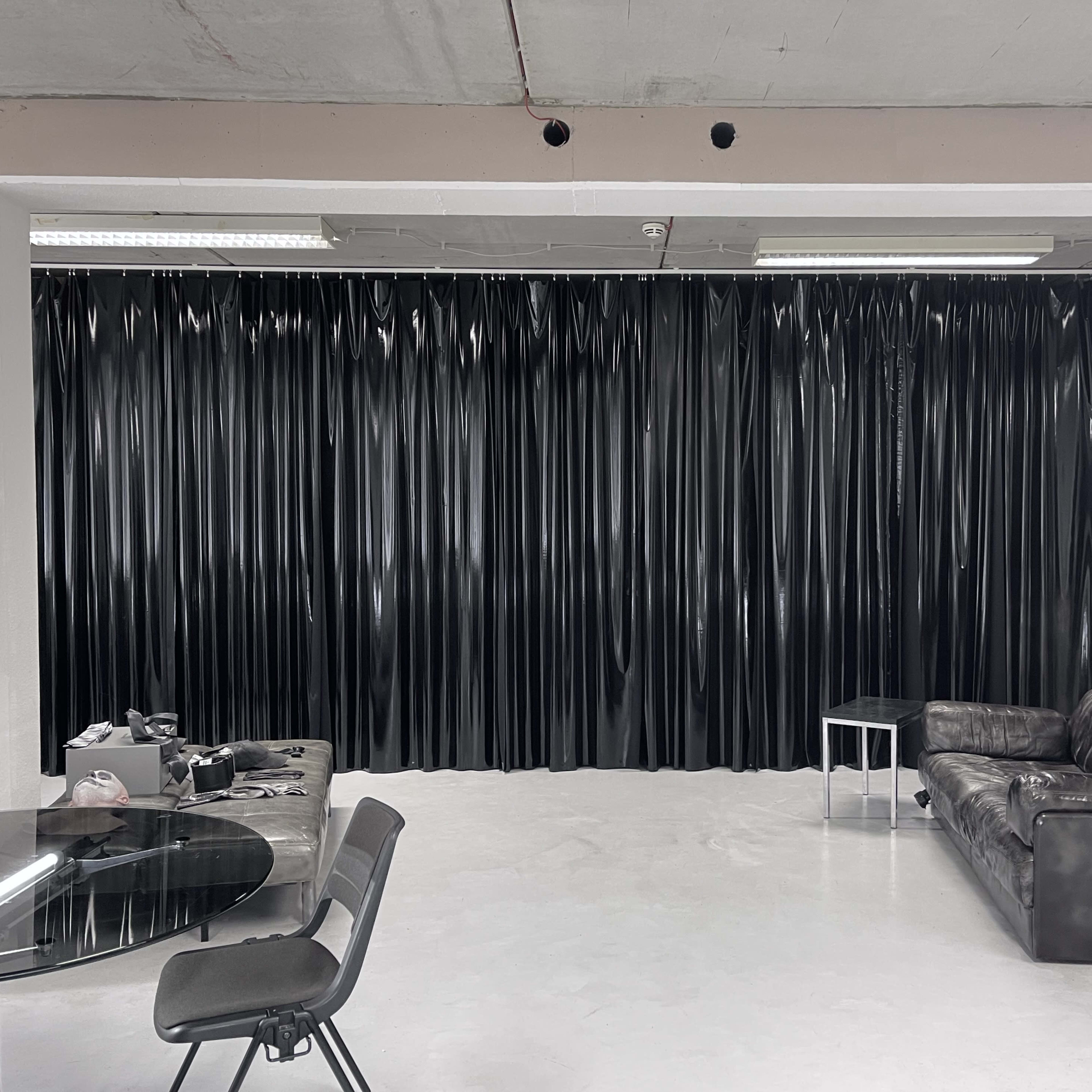 A black curtain lines the wall of a minimalistic room featuring a glass dining table, several chairs, and a leather sofa.