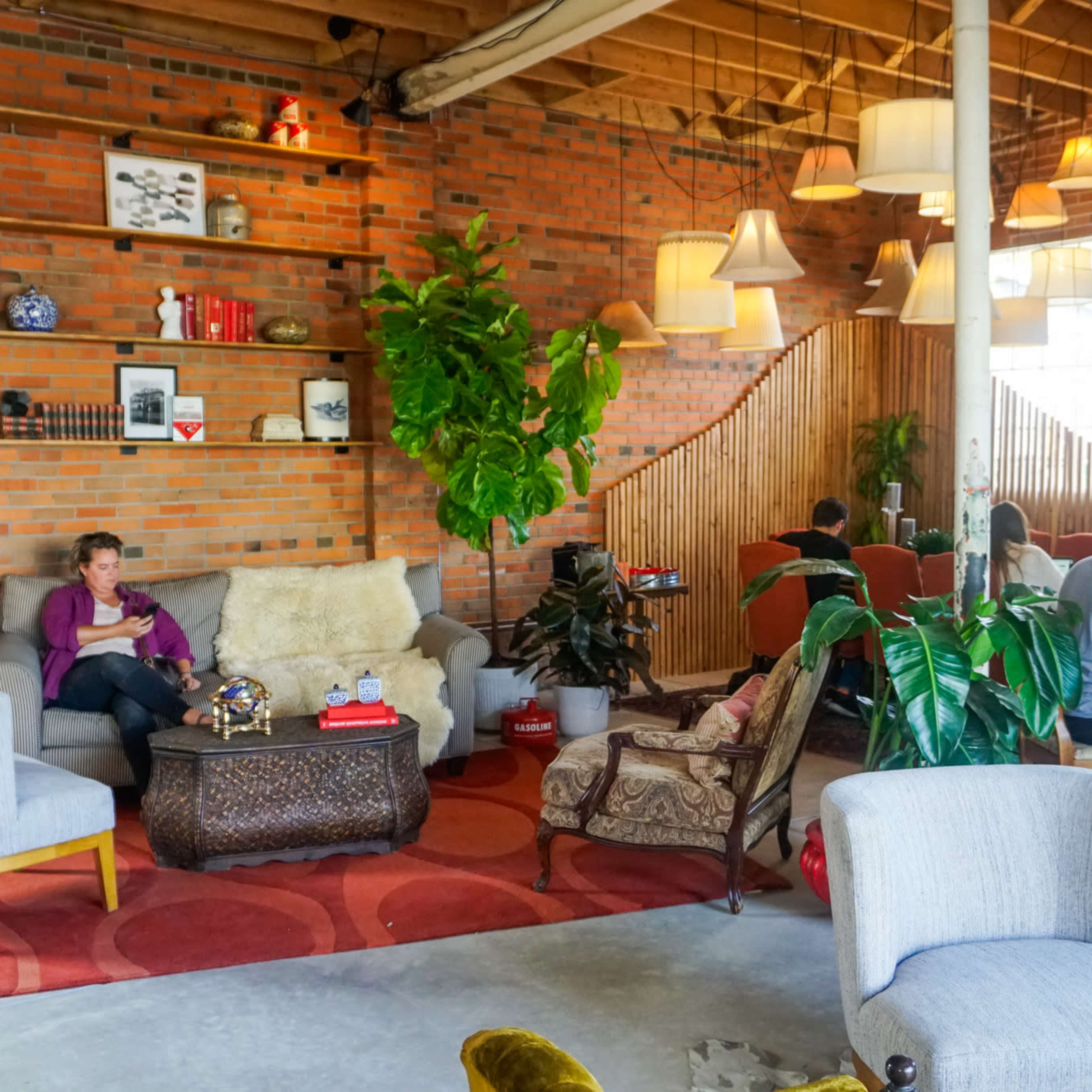 A cozy, modern cafe features a mix of seating areas, including a woman reading on a sofa, while others work on laptops at tables, with plants and decorative lighting enhancing the ambiance.