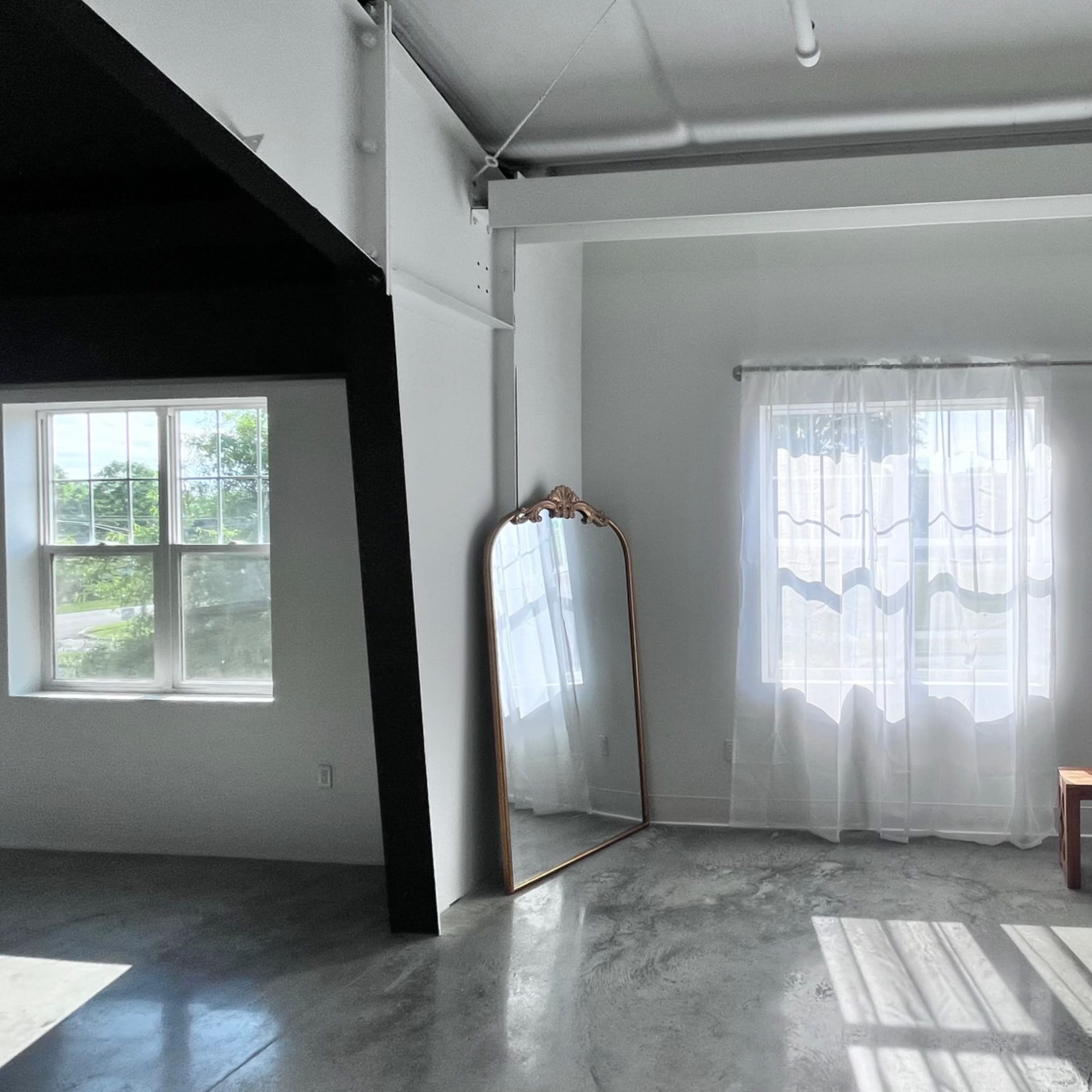 A room with two large windows covered by sheer curtains, a wooden bench in the center, and a tall mirror leaning against the wall.