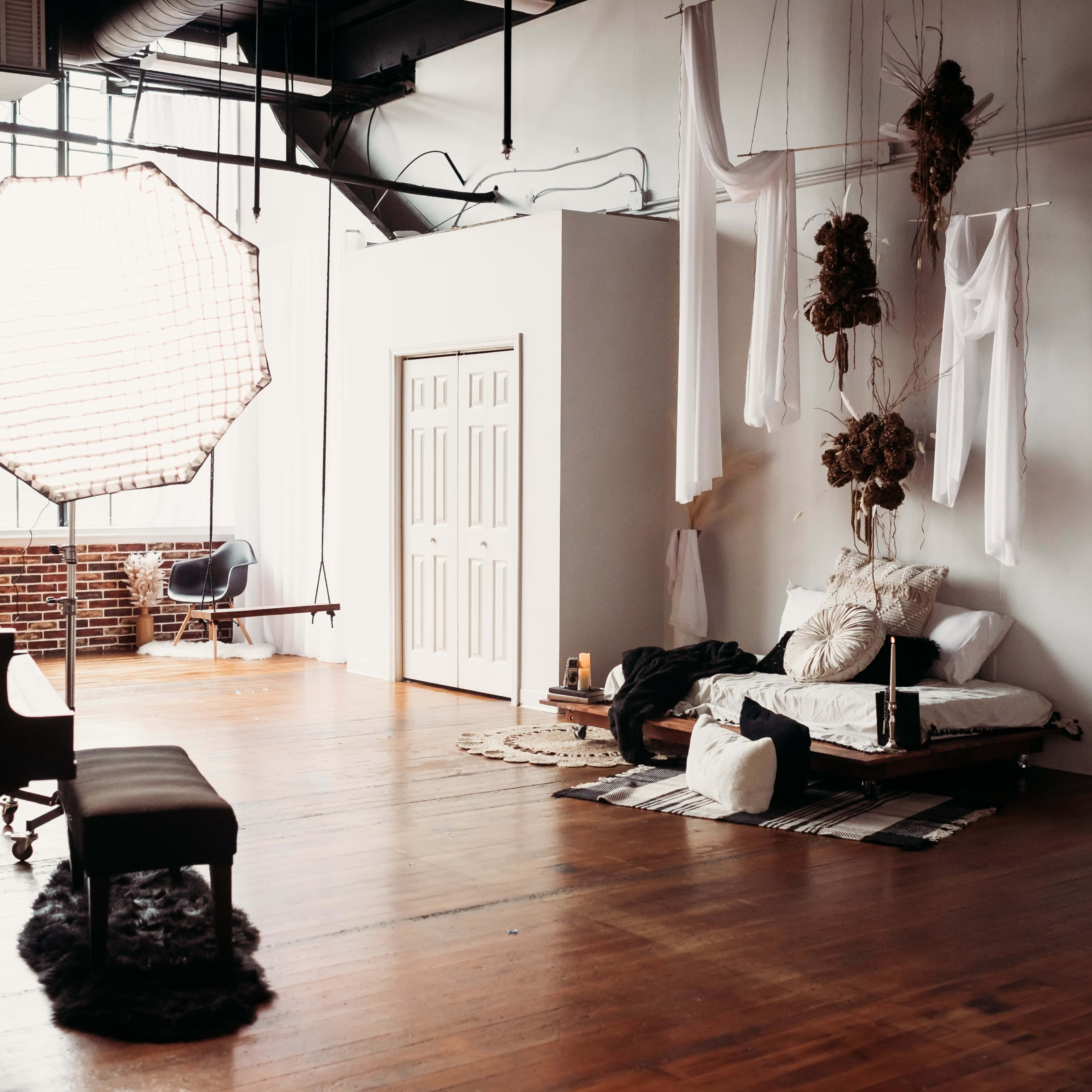 A spacious studio features a grand piano, a bed adorned with various textiles, and soft lighting from a large diffuser near a large window.