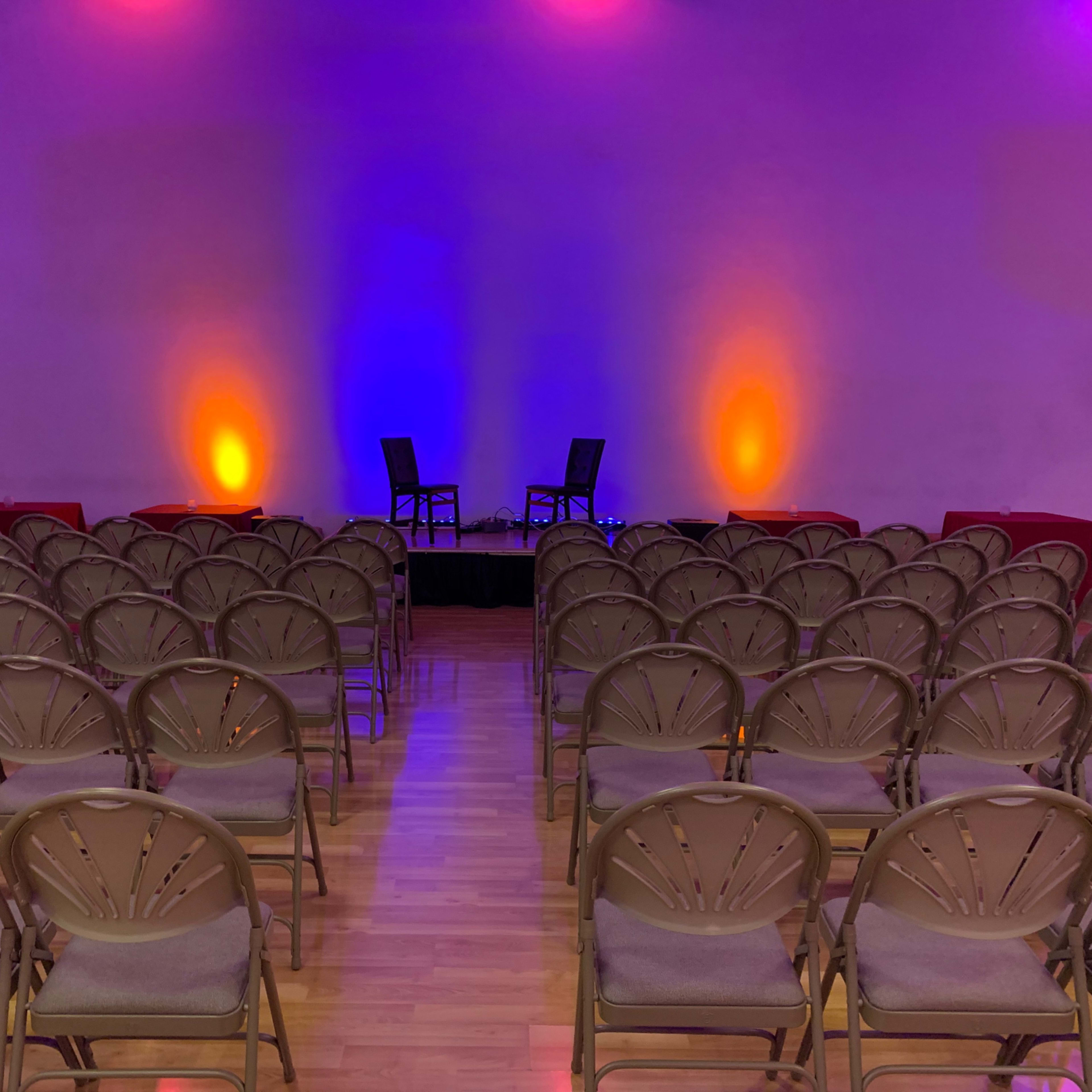 A room set up for an event with rows of empty chairs facing a stage flanked by two illuminated chairs.