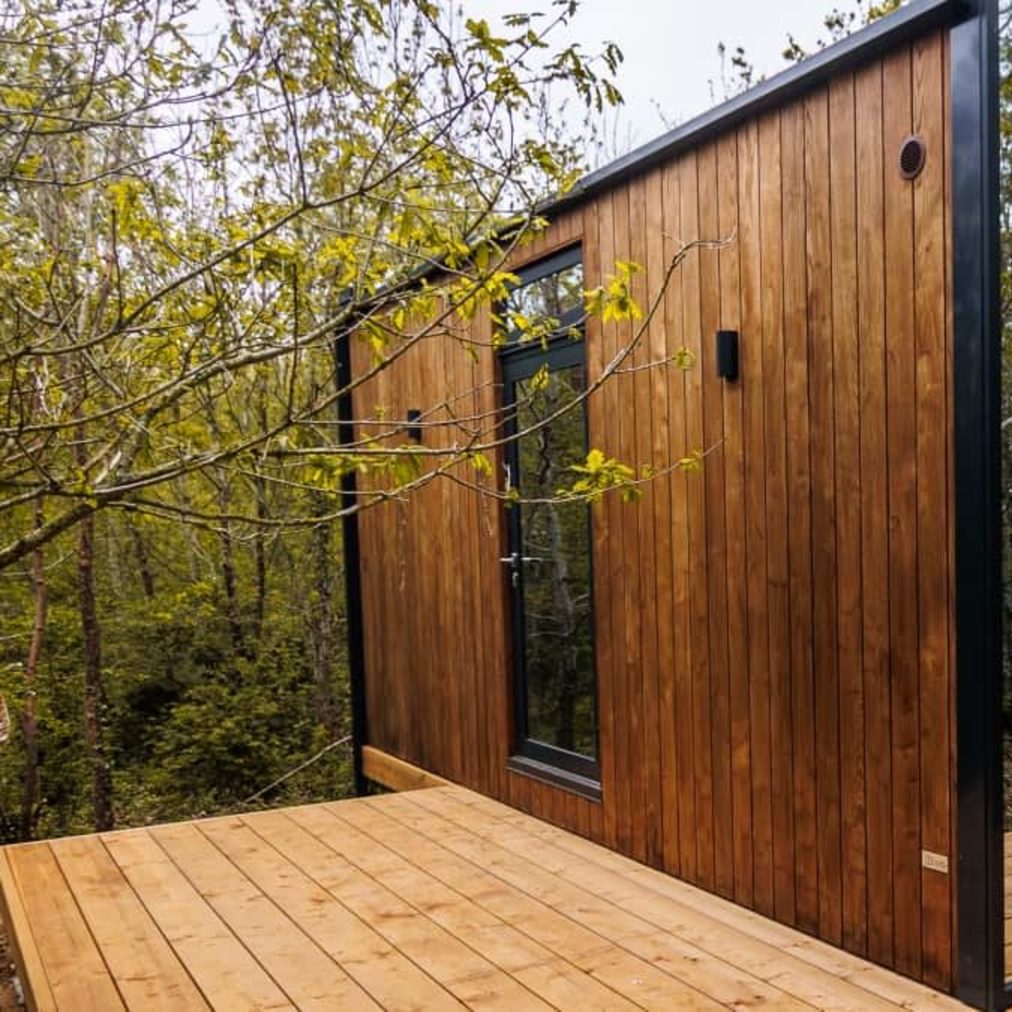 A modern wooden cabin with one side featuring large mirrored glass panels, surrounded by trees and set on a wooden deck.
