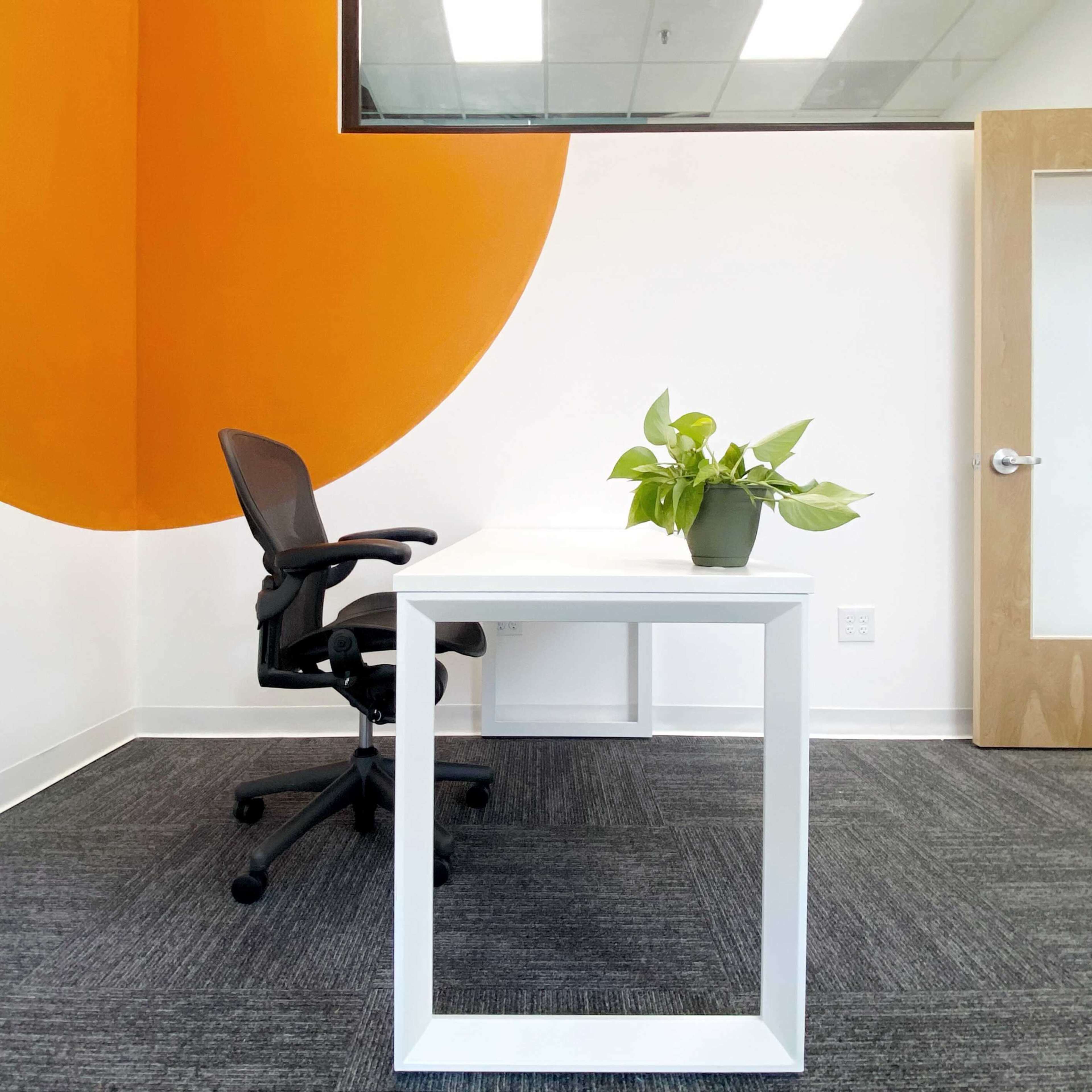 A minimalist office features a white desk and a black chair beside a potted plant, with an orange accent wall and a door in the background.